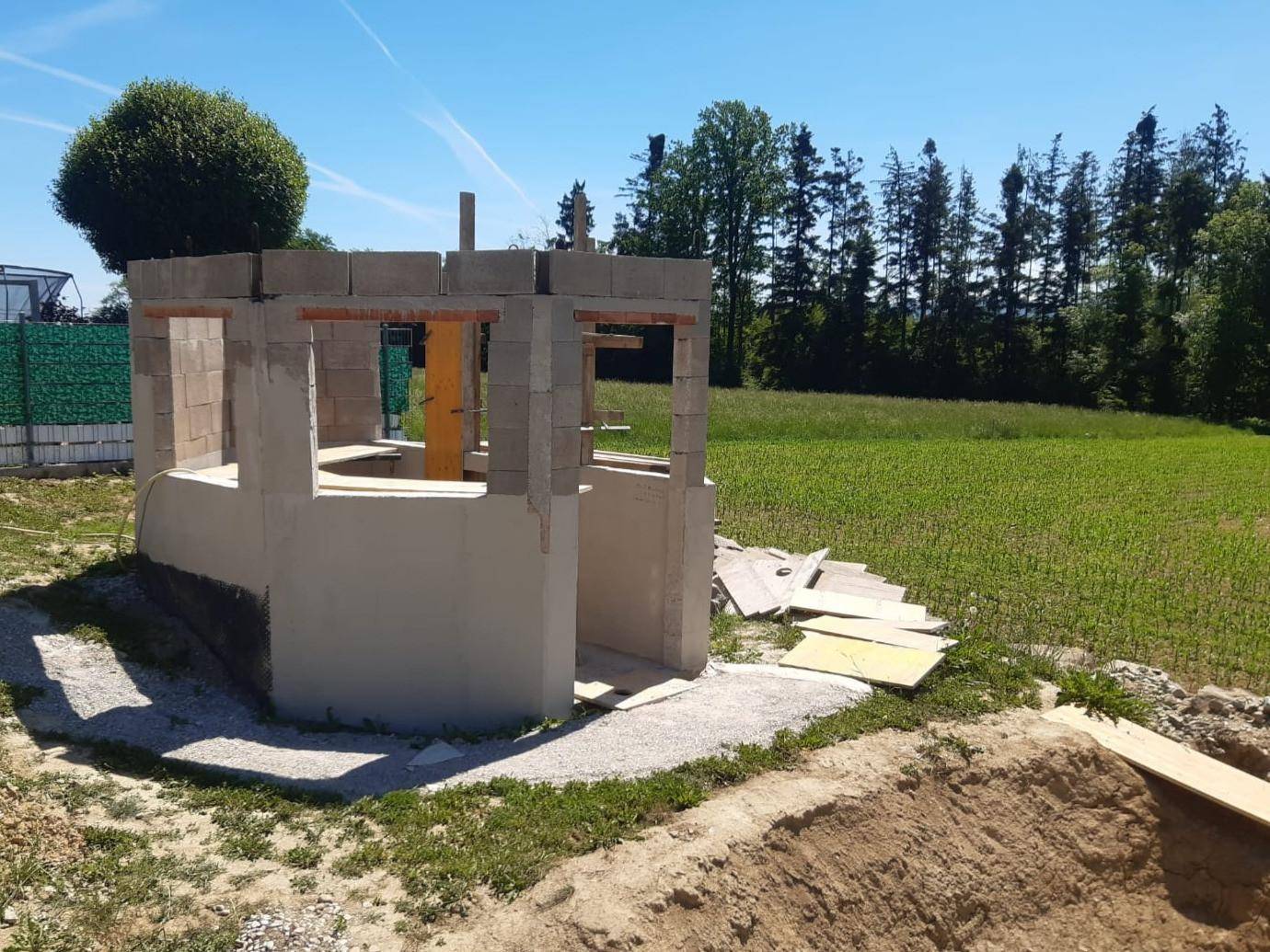 Baustelle für eine zukünftige Außenstruktur mit Blick auf grüne Felder und Bäume im Hintergrund.