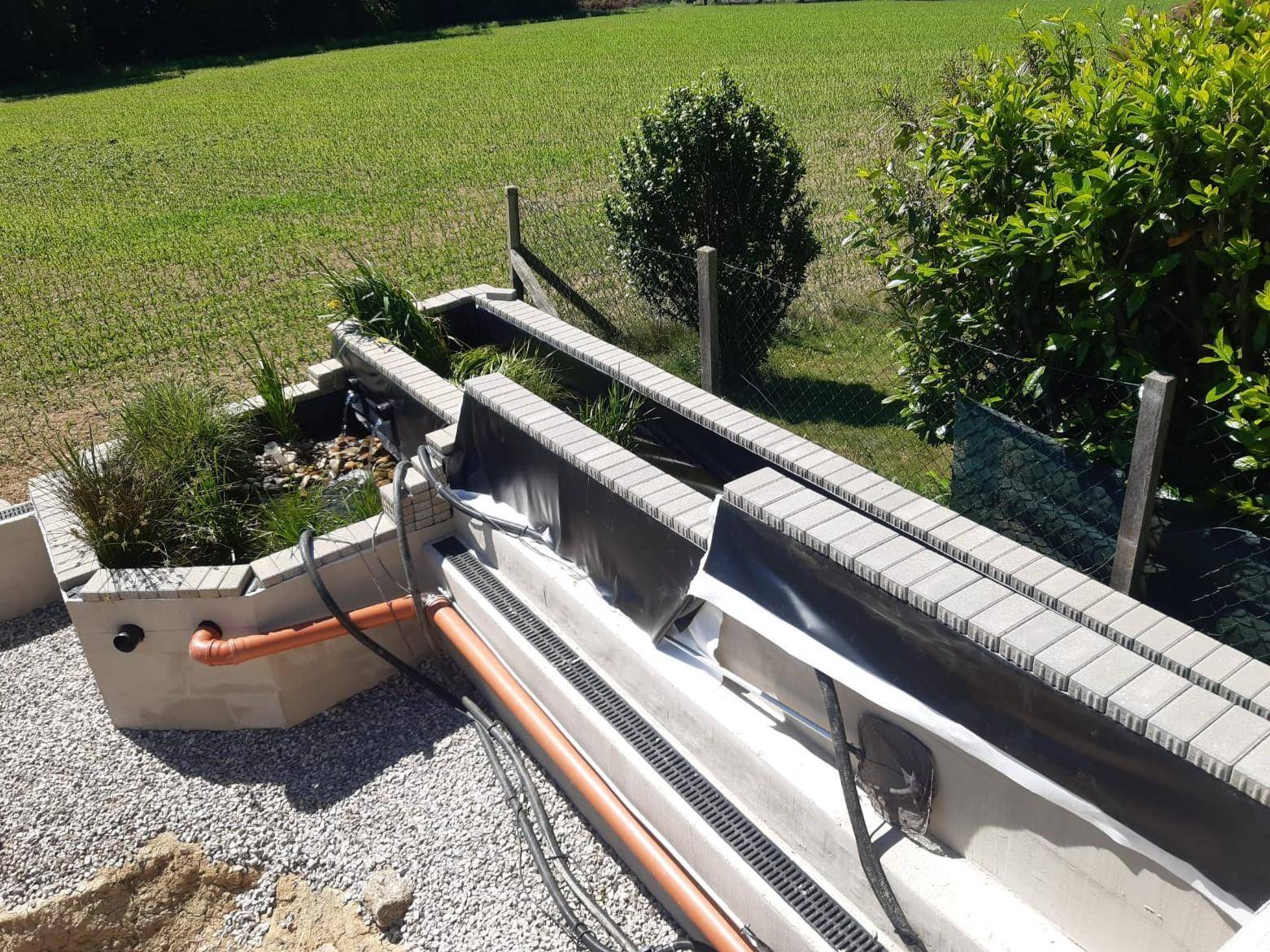 Gartenbereich mit im Bau befindlichem Wasserbecken und Bepflanzung, umgeben von Grünflächen.