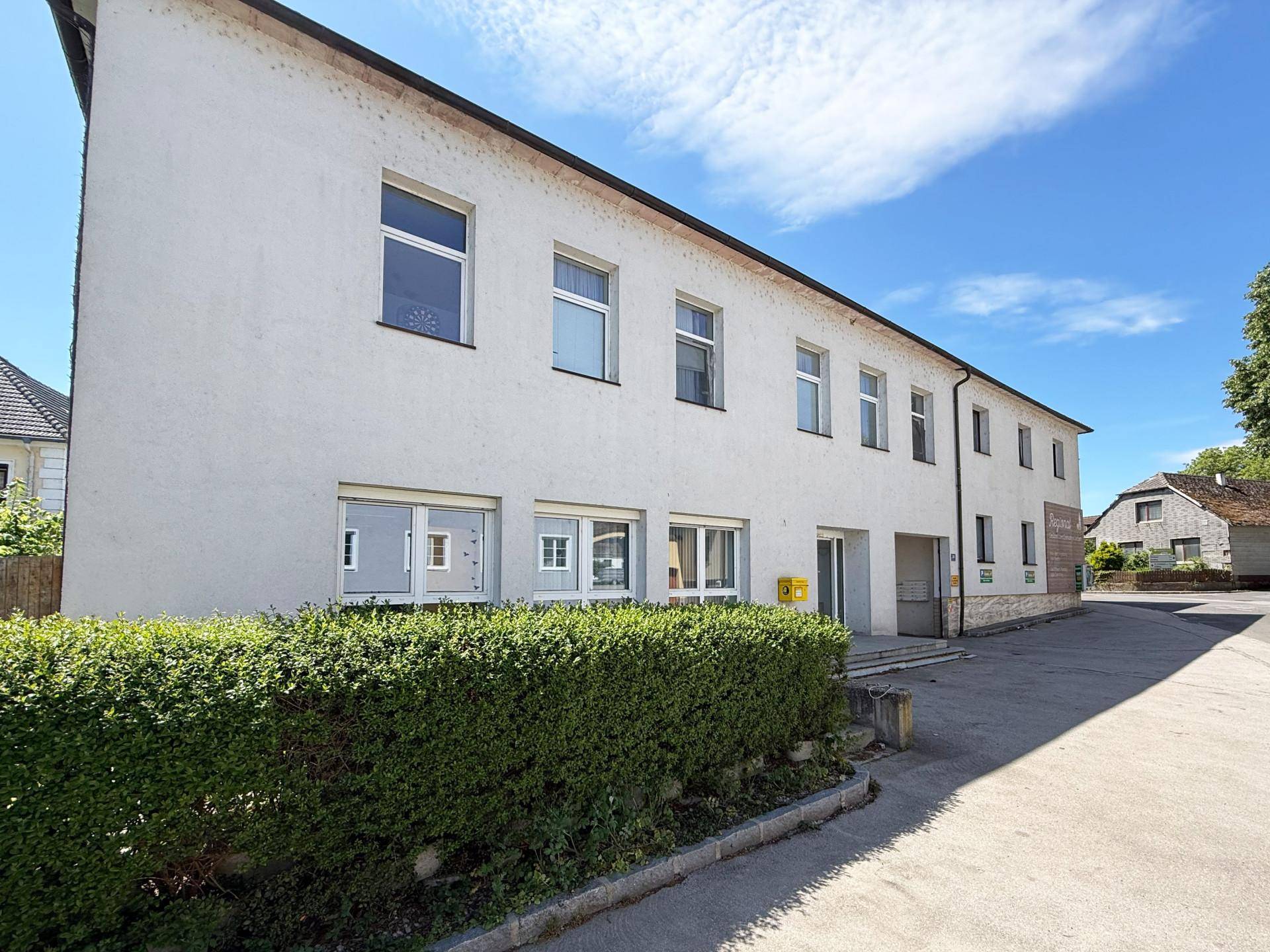Seitenansicht eines weißen Gebäudes mit vielen Fenstern und einer gepflegten Hecke im Vordergrund.