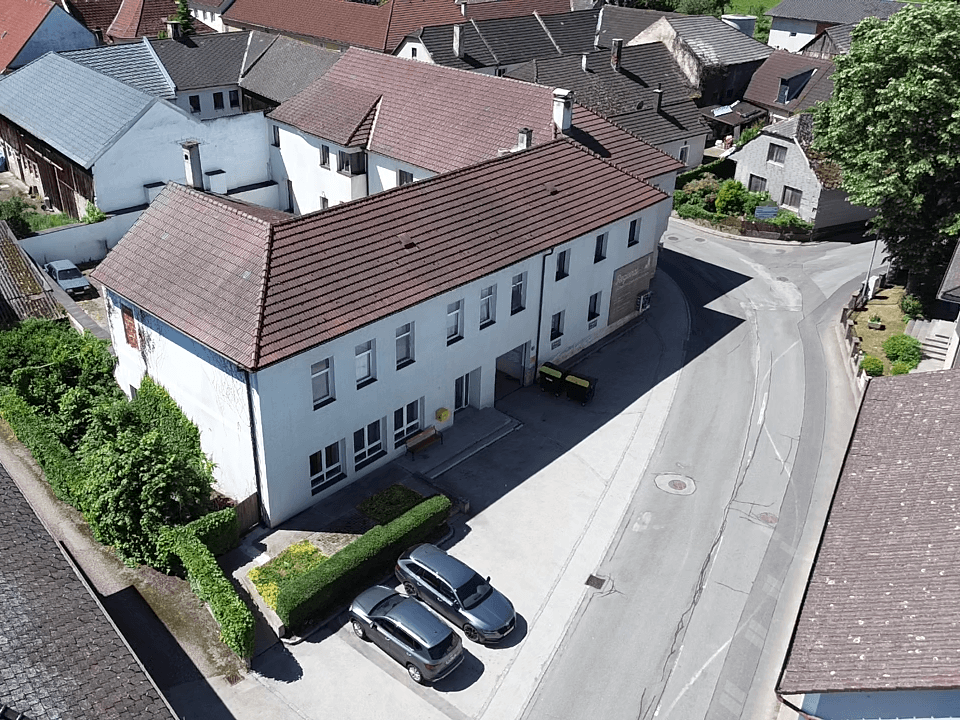 Luftaufnahme eines langgestreckten Gebäudes mit rotem Ziegeldach, Parkplätzen und einer Straße daneben.
