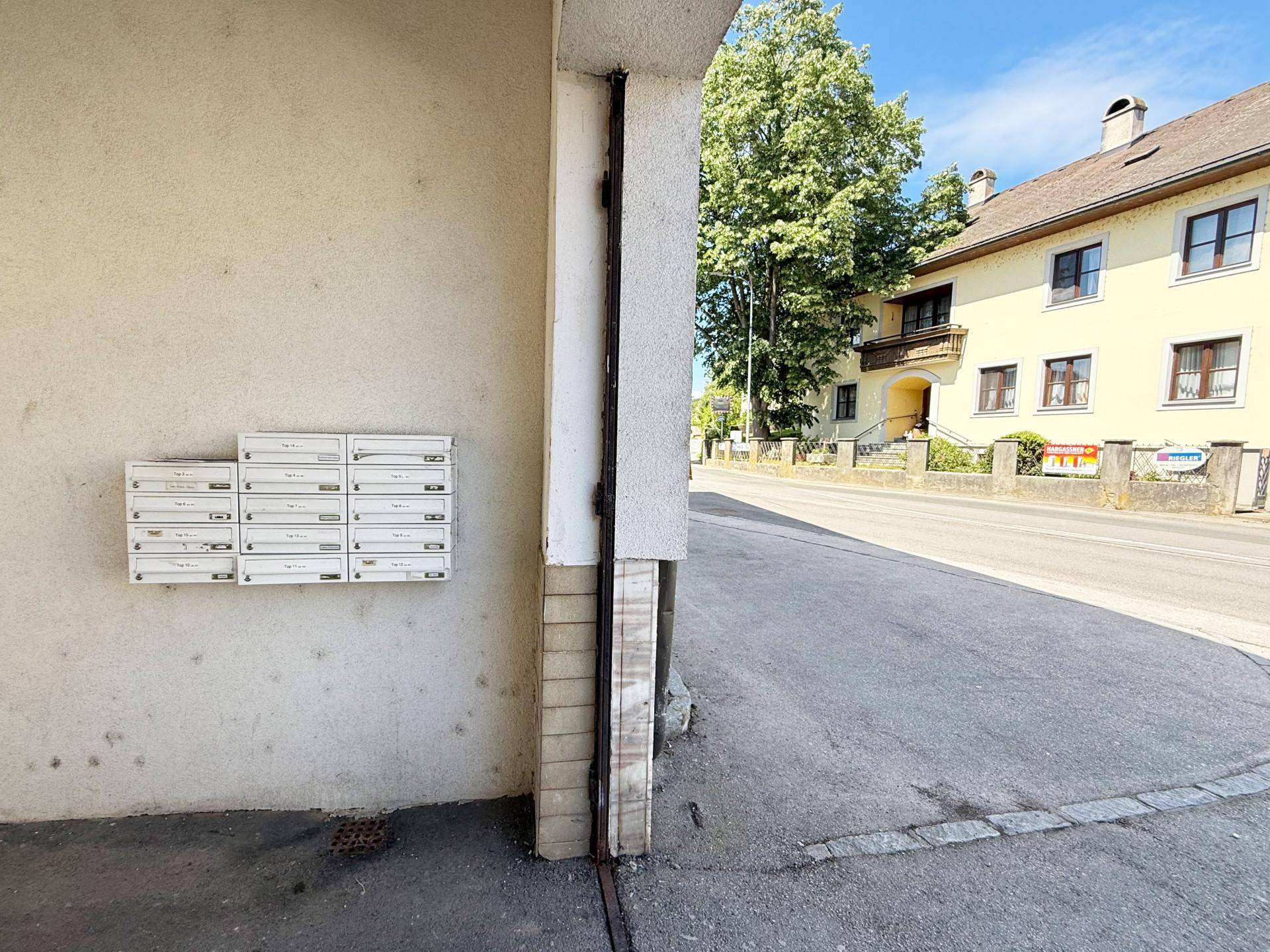 Außenansicht mit mehreren Briefkästen an einer Gebäudewand und einer Straße mit Bäumen im Hintergrund.