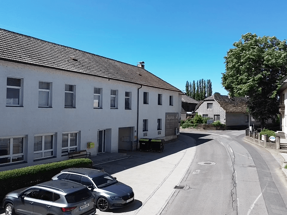 Luftaufnahme eines Gebäudes mit rotem Ziegeldach, Parkplätzen und einer Straße, umgeben von Grünflächen.