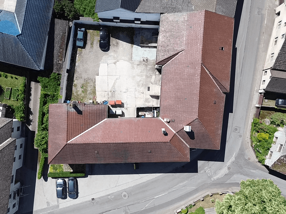Senkrechte Luftaufnahme eines L-förmigen Gebäudes mit roten Ziegeldächern und einem Innenhof mit Parkplätzen.