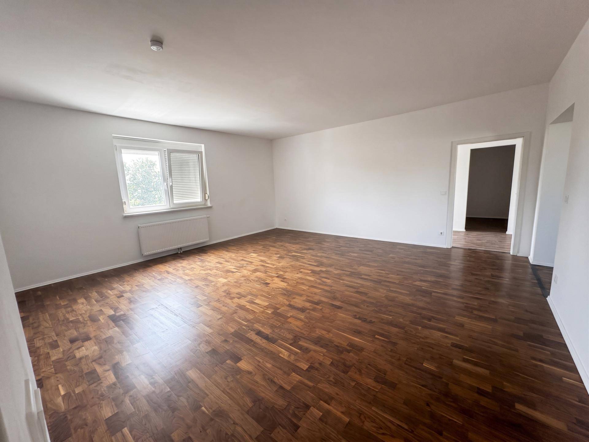 Leeres Wohnzimmer mit dunklem Parkettboden, weißen Wänden und einem Fenster.