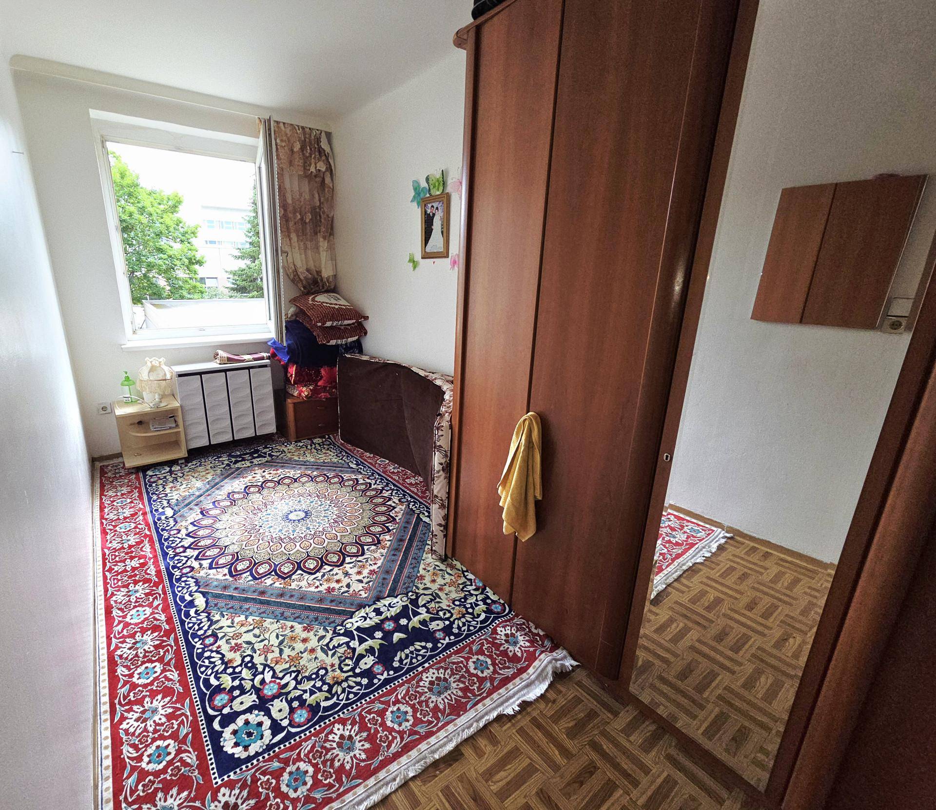 Helles Schlafzimmer mit gemustertem Teppich, Holzschrank und Blick ins Grüne.