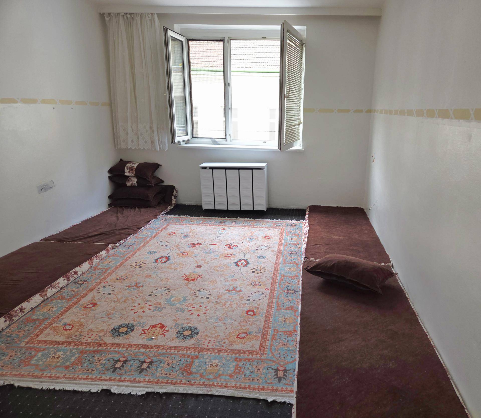 Helles Zimmer mit gemustertem Teppich, Heizkörper unter dem Fenster und Blick nach draußen.
