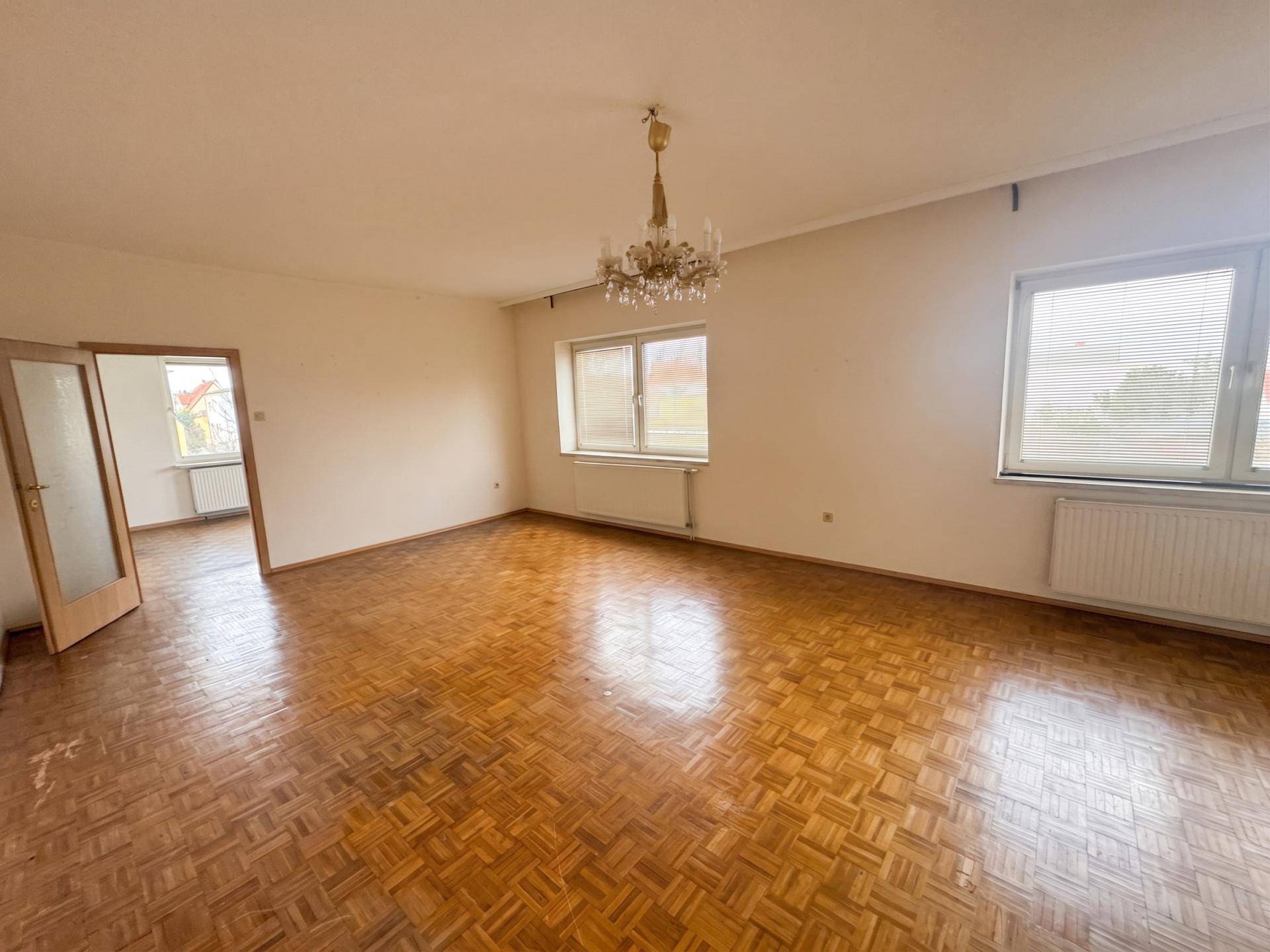 Geräumiges, helles Wohnzimmer mit Parkettboden, weißen Wänden und einem eleganten Kronleuchter.
