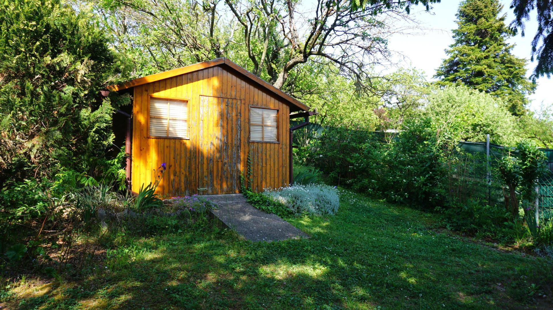 Kleiner Holzschuppen im grünen Garten, umgeben von Bäumen und Sträuchern an einem sonnigen Tag.