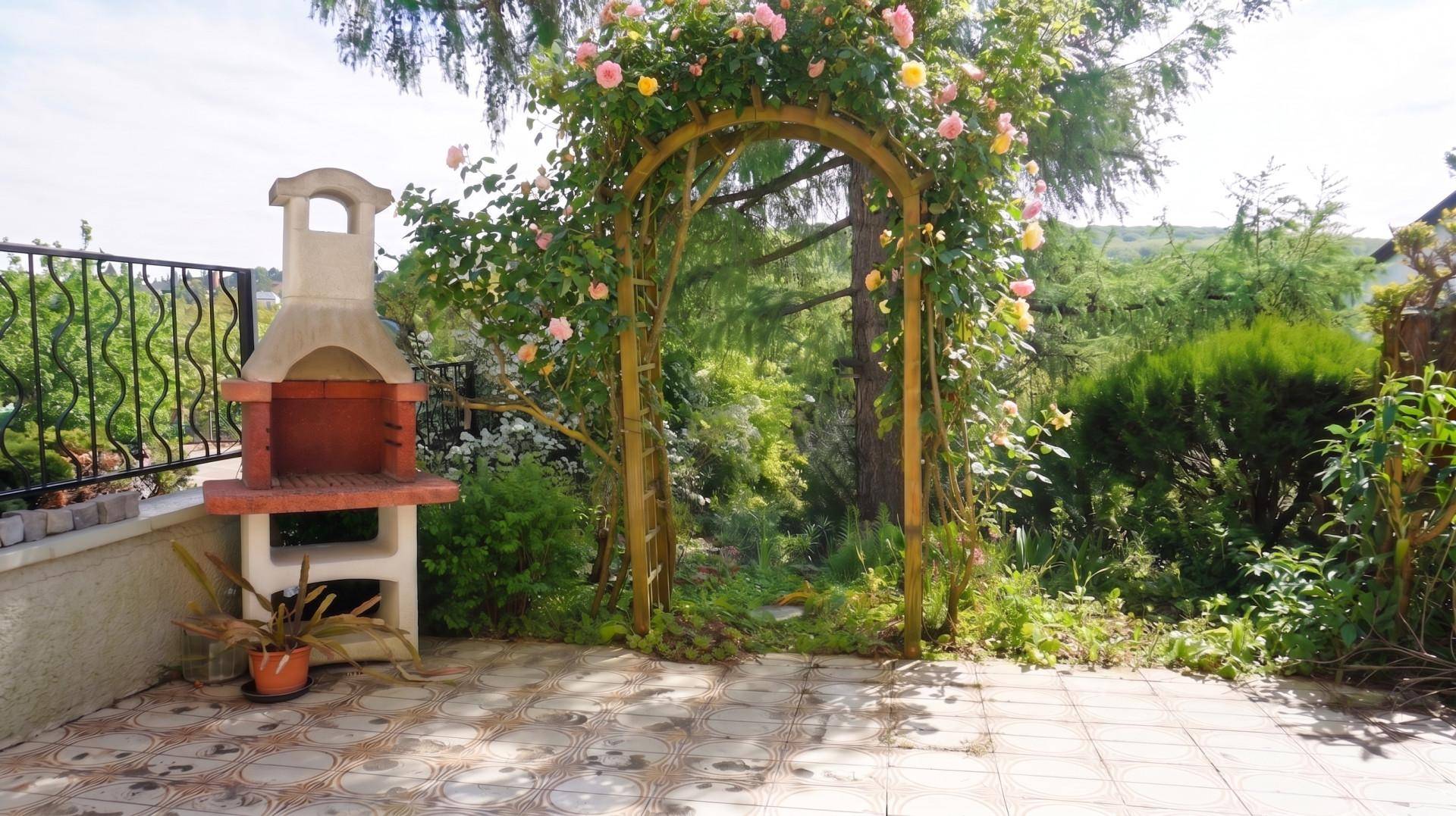 Geflieste Terrasse mit gemauertem Grill und blühendem Rosenbogen, umgeben von üppigem Grün.