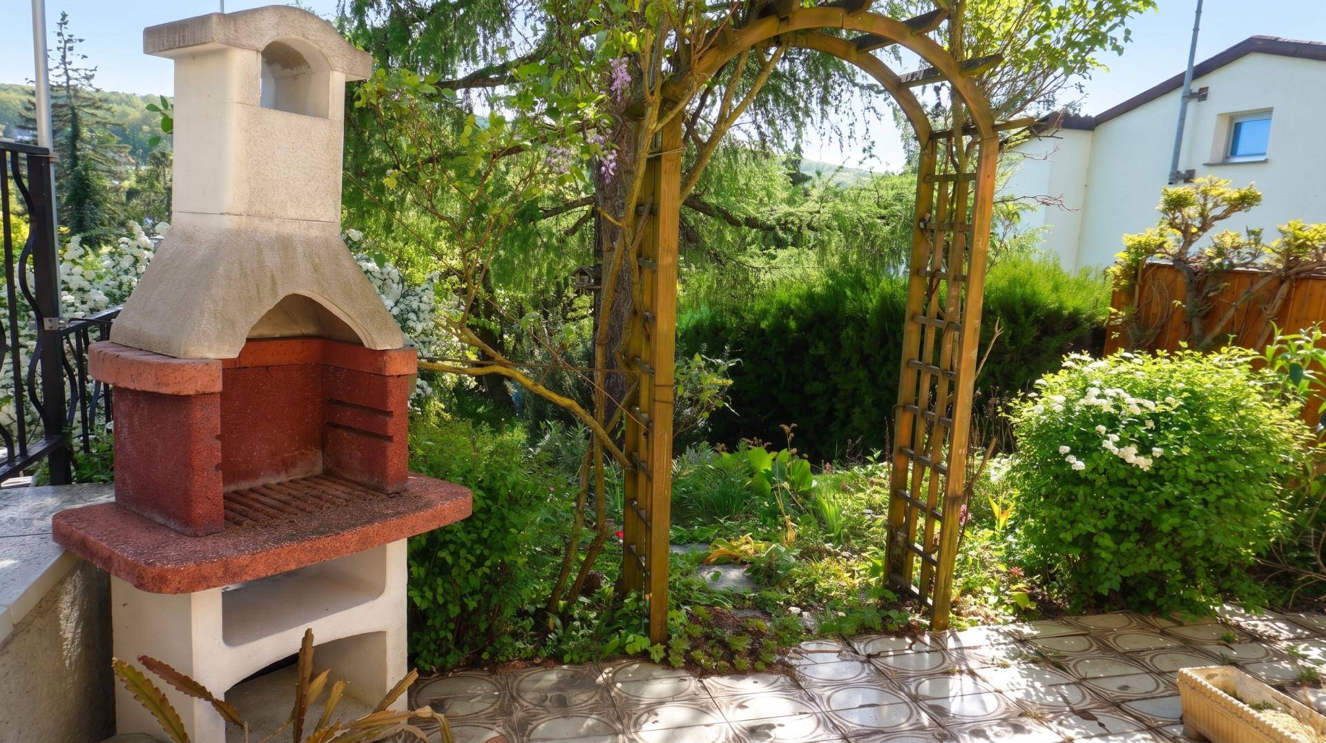 Geflieste Terrasse mit gemauertem Grill und blühendem Rosenbogen, umgeben von üppigem Grün.
