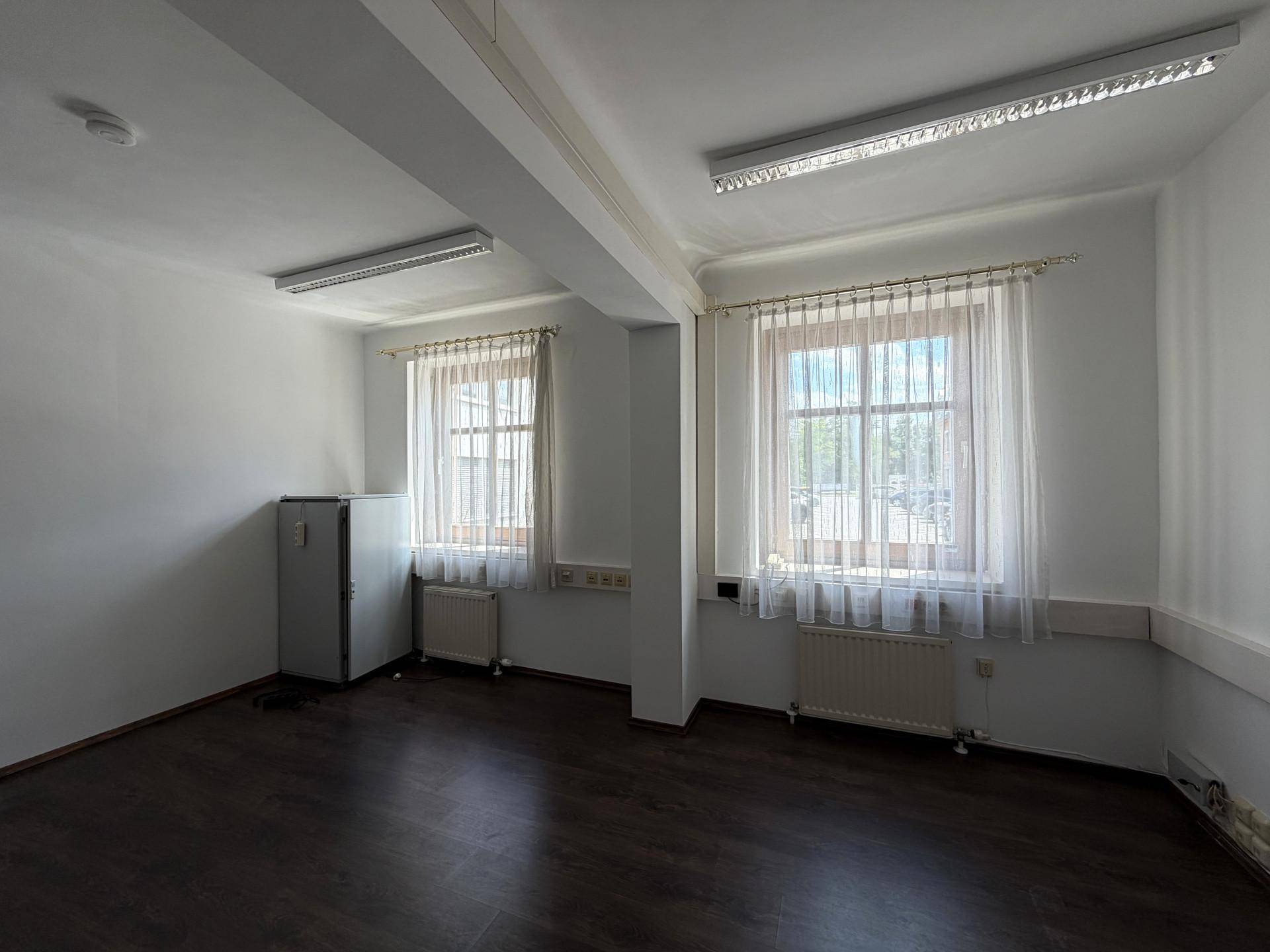 Geräumiger Raum mit zwei Fenstern, dunklem Boden und einem Kühlschrank, ideal als Pausenraum.