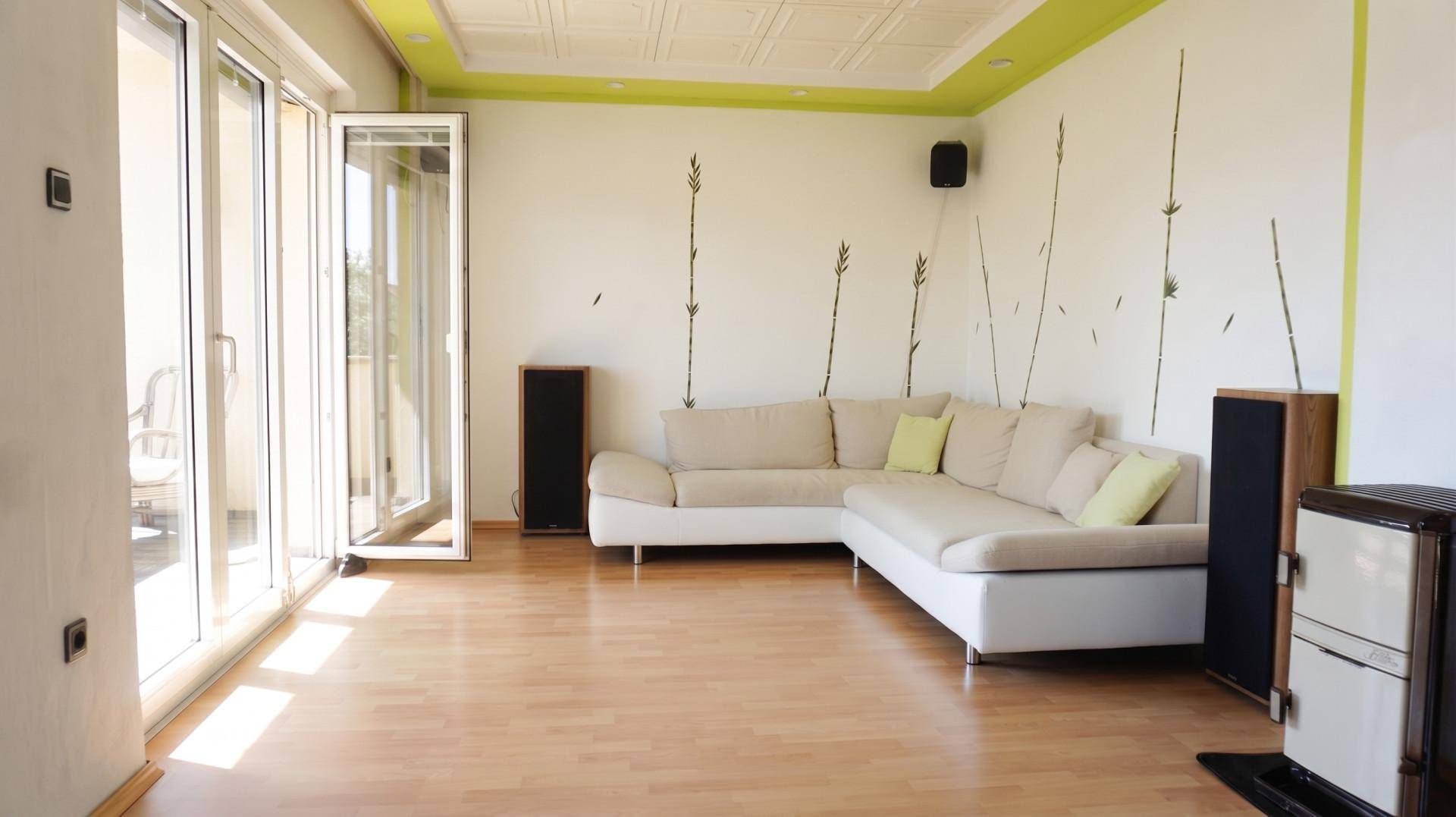 Helles Wohnzimmer mit Laminatboden, Ecksofa und Zugang zum Balkon durch große Fenster.