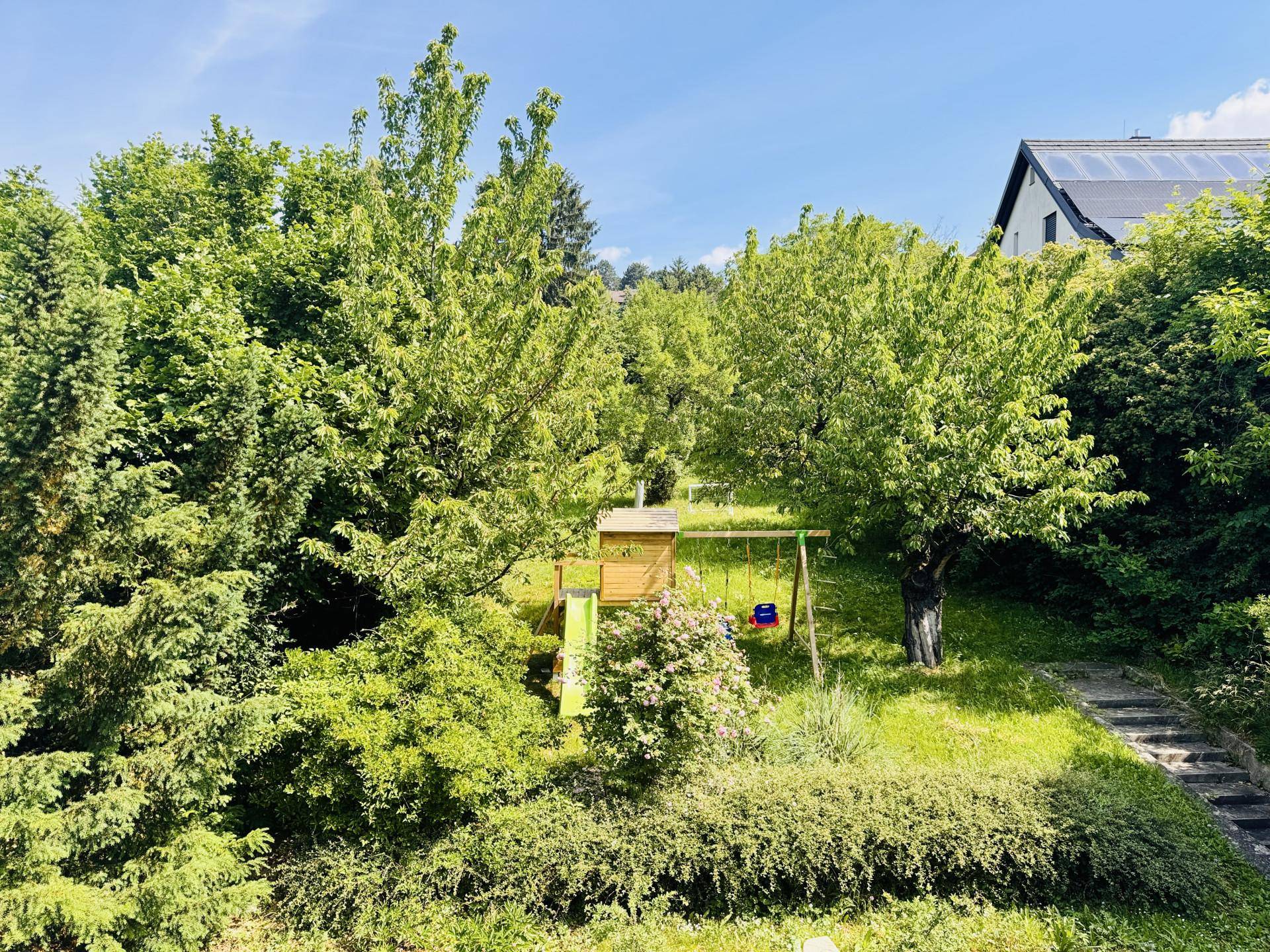 Weitläufiger Garten mit Spielplatz, Schaukel und Rutsche, umgeben von dichtem Baumbestand und Rasenfläche.