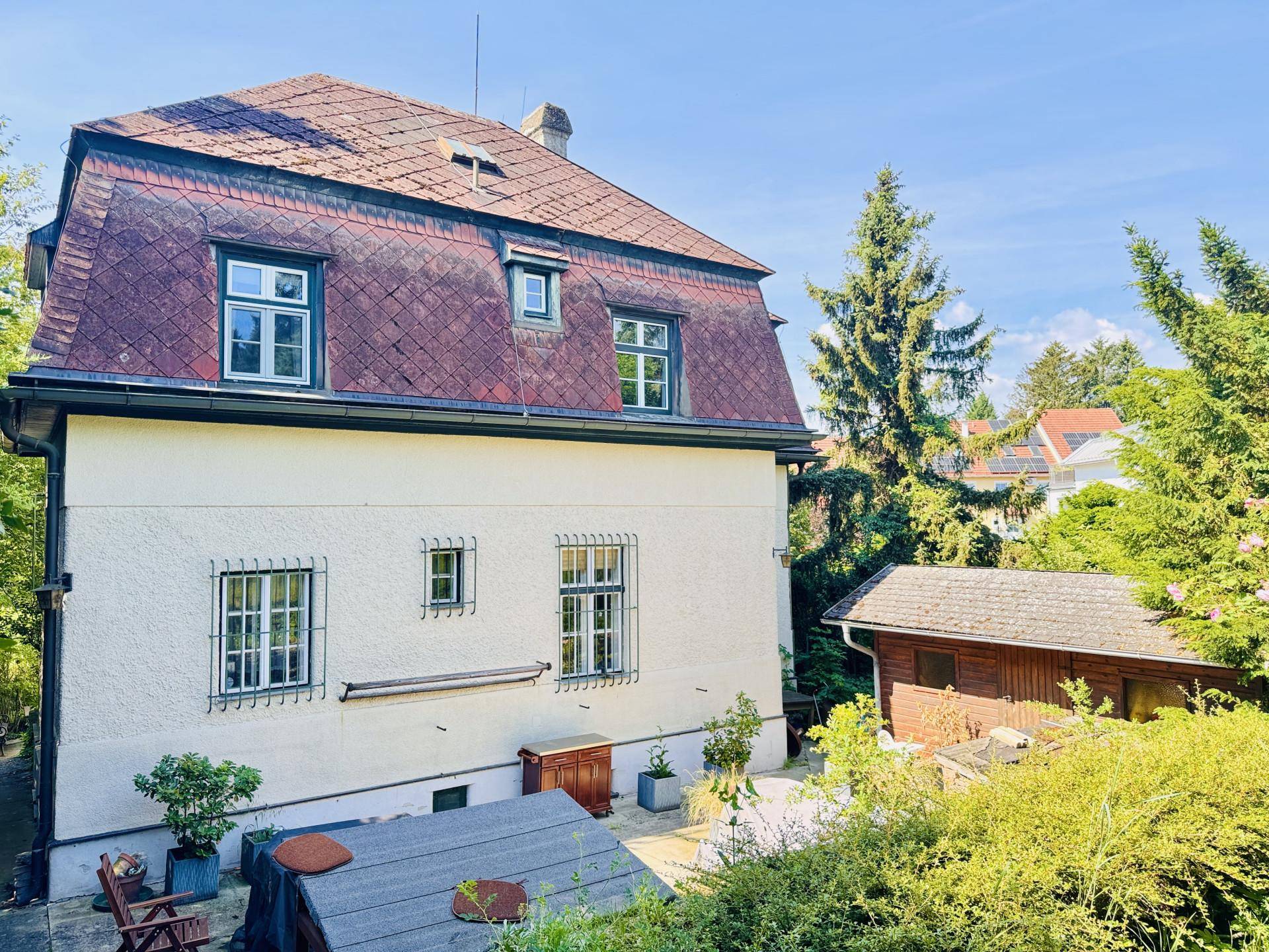 Rückansicht des Hauses mit Mansarddach und einem kleinen Gartenhaus, umgeben von Bäumen und Sträuchern.