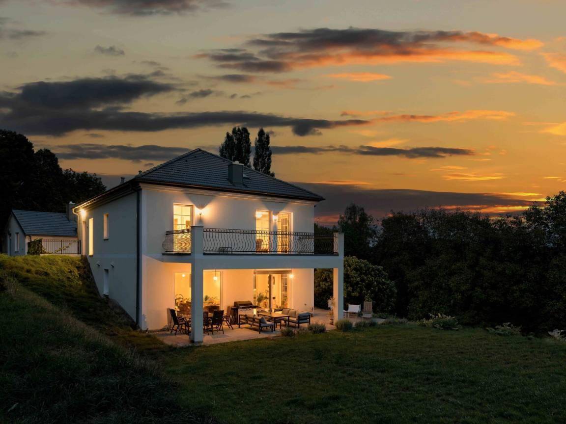 Das Haus bei Sonnenuntergang, mit beleuchteten Innenräumen und stimmungsvollem Himmel.
