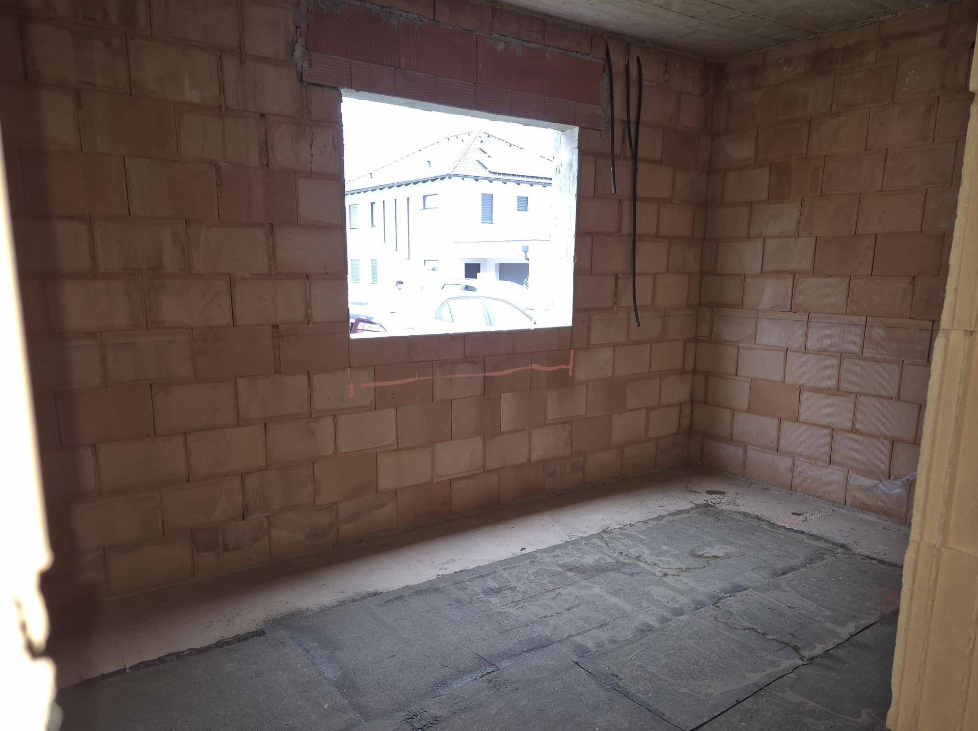 Rohbauzimmer mit Ziegelwänden, Betonboden und einer Fensteröffnung mit Blick auf ein Nachbargebäude.