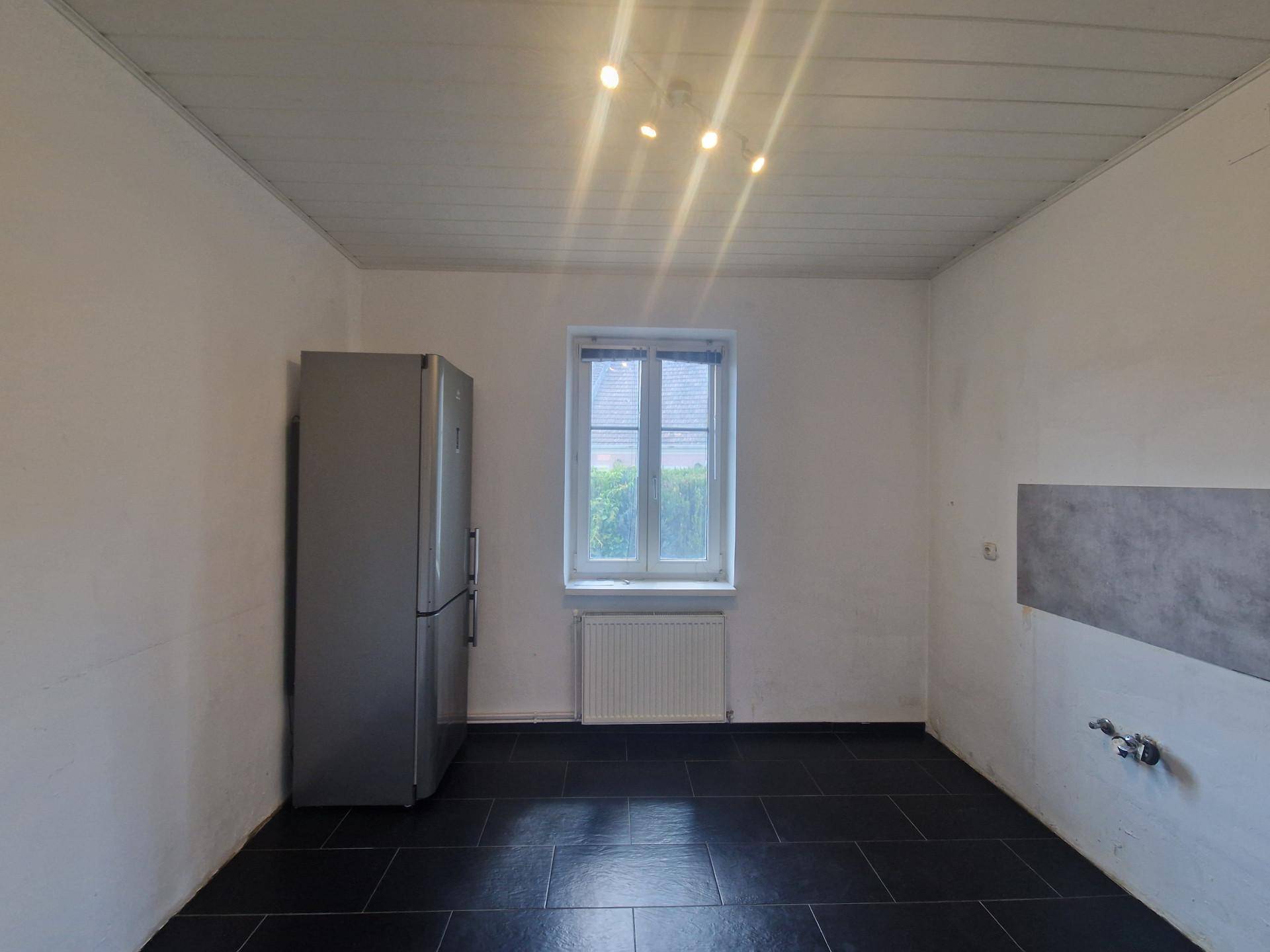 Leere Küche mit schwarzem Fliesenboden, Kühlschrank und Fenster mit Blick ins Grüne.