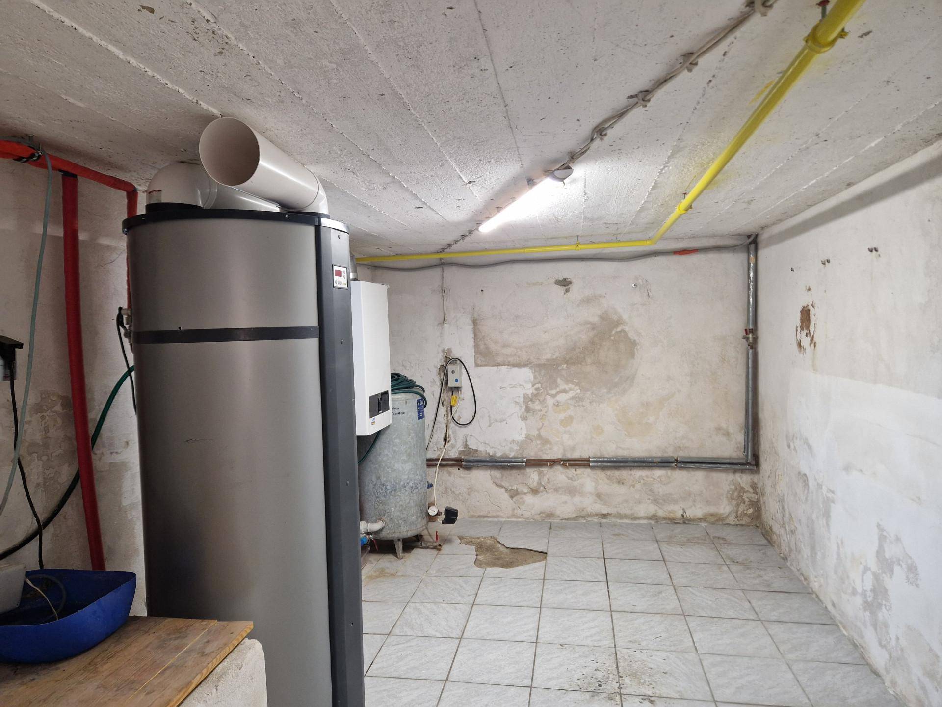 Kellerraum mit Warmwasserspeicher, sichtbaren Leitungen und Fliesenboden.