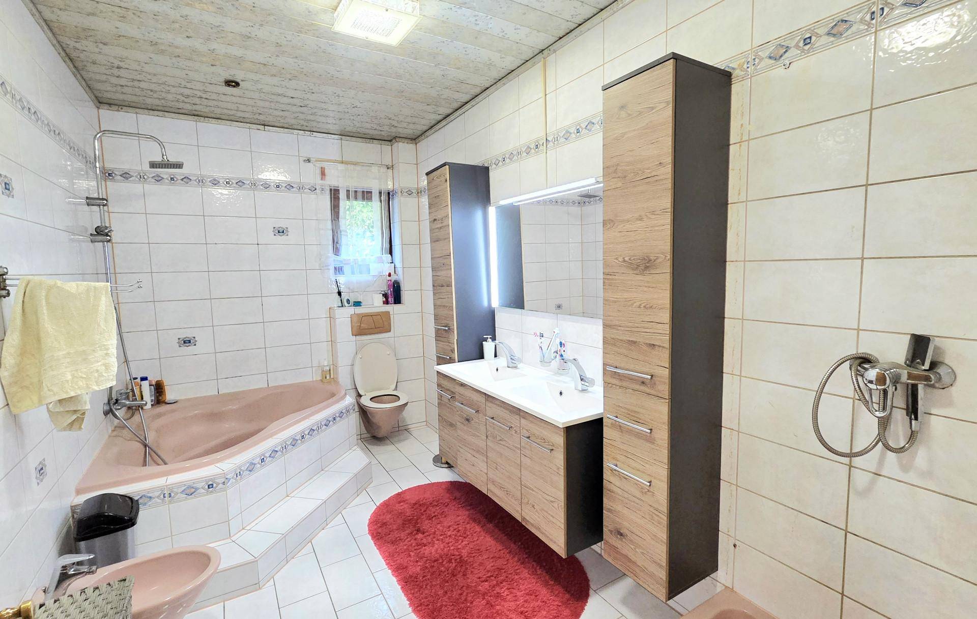 Badezimmer mit rosa Eckbadewanne, Doppelwaschbecken und Holzschränken, gefliest in Weiß und Braun.