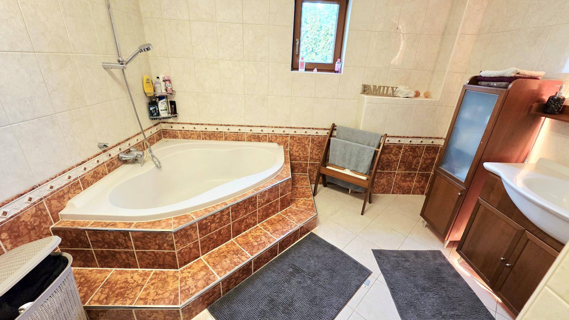 Badezimmer mit großer Eckbadewanne, braunen Fliesen und Holzschrank, gut beleuchtet durch ein Fenster.