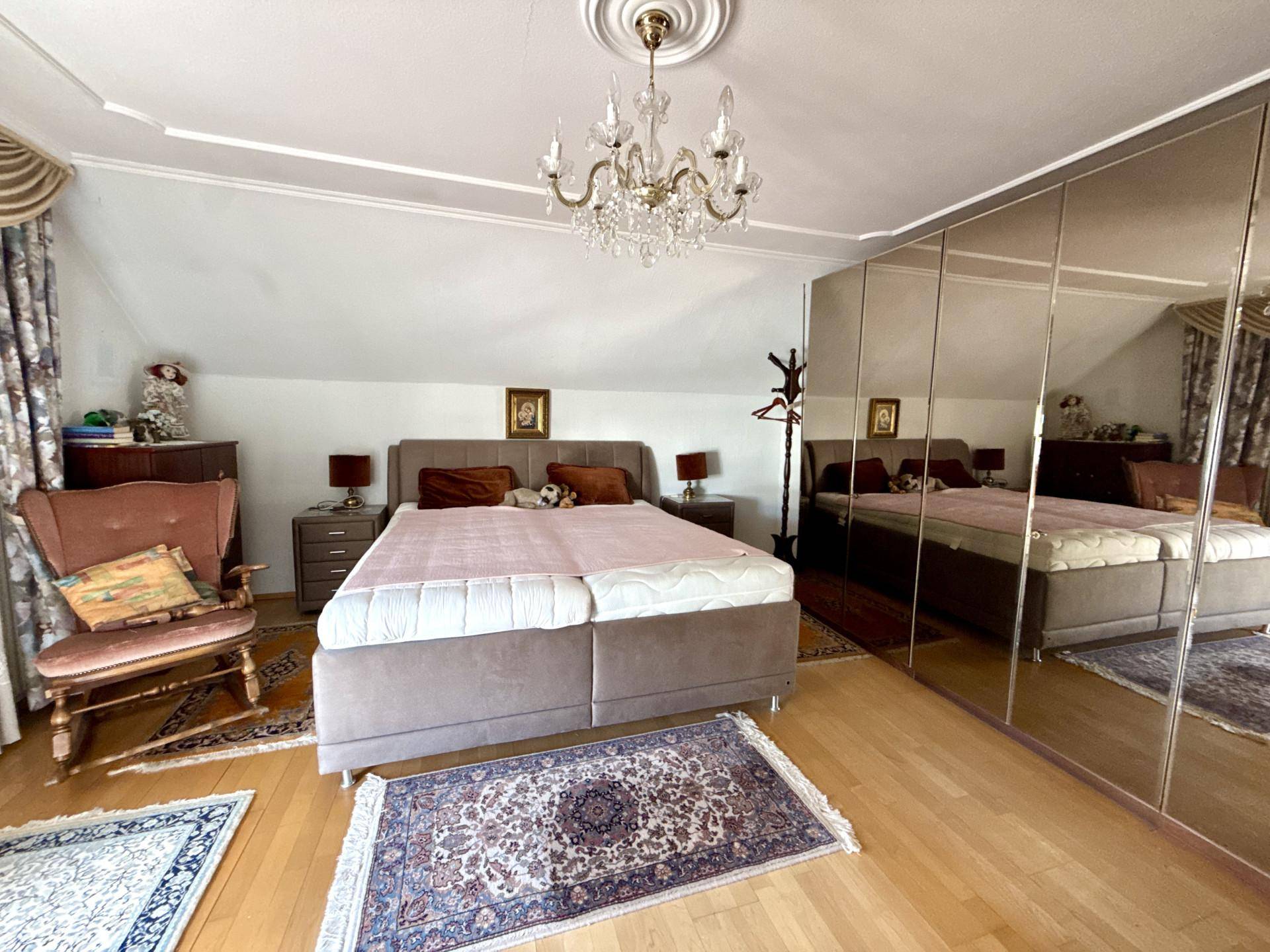 Helles Schlafzimmer mit Doppelbett, Spiegelschrank und Dachschräge unter einem Kronleuchter.