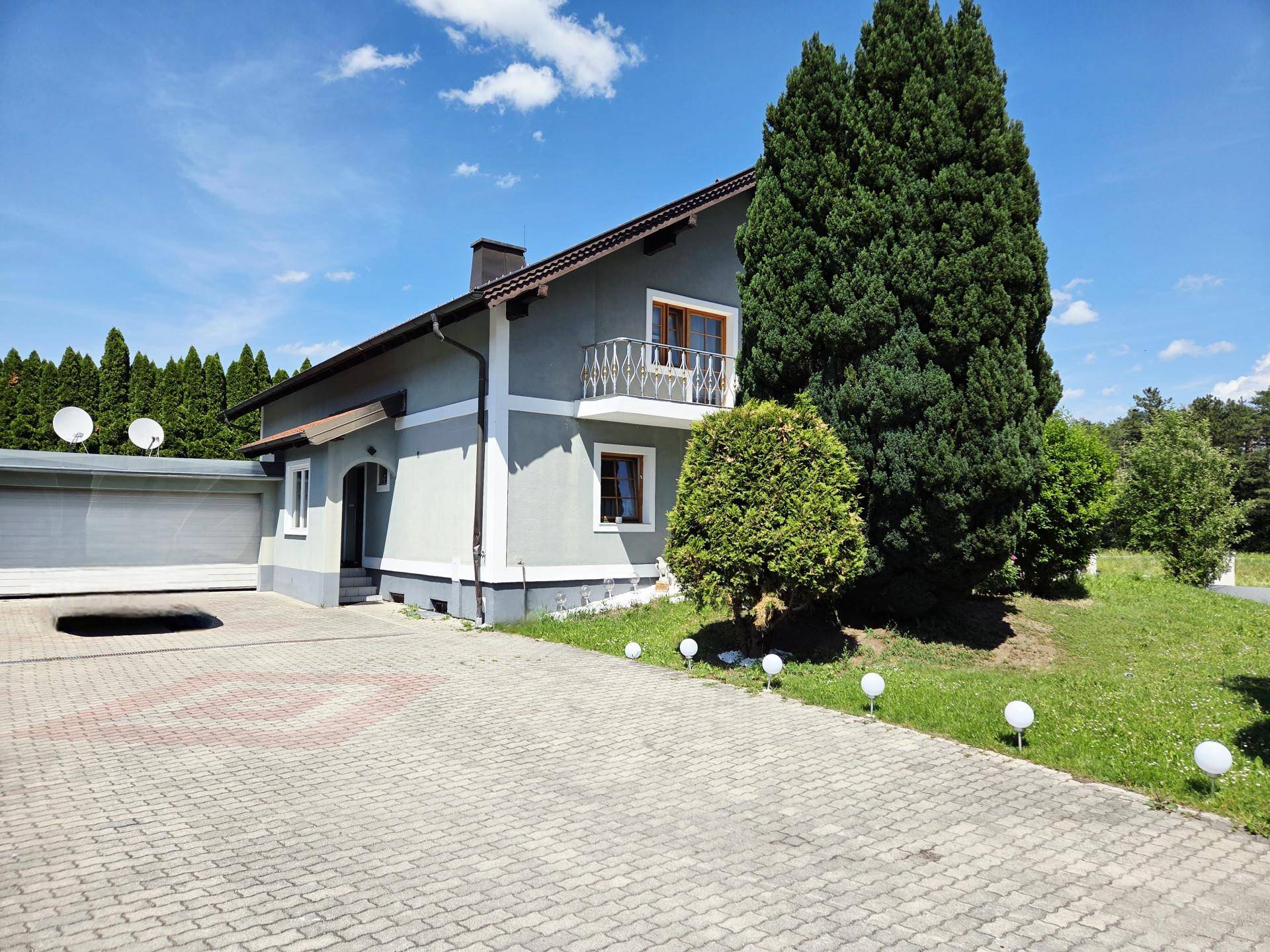 Seitenansicht des Hauses mit gepflasterter Auffahrt, Garage und hohen Bäumen im Hintergrund.