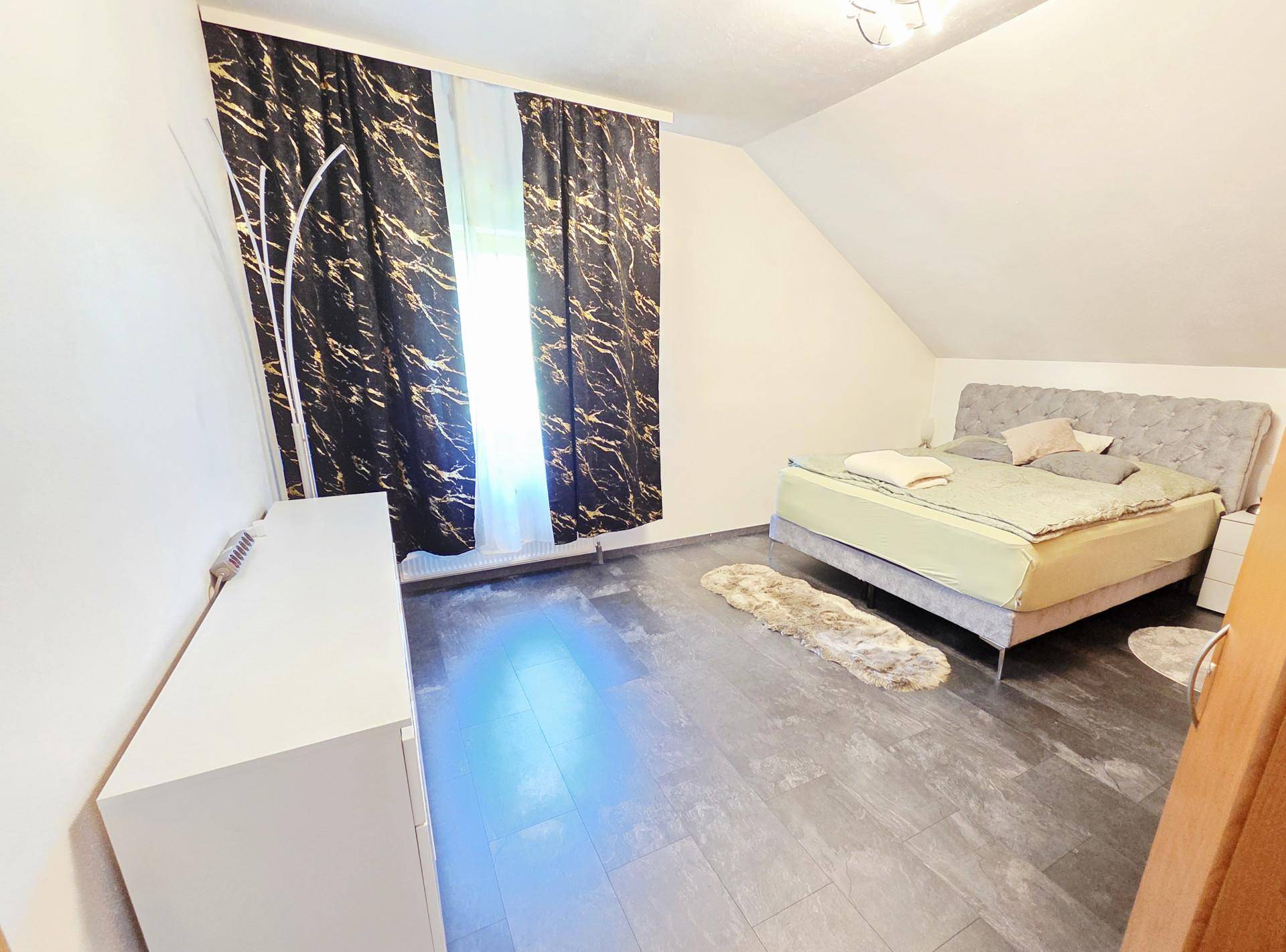 Schlafzimmer im Dachgeschoss mit modernem Bett, dunklem Boden und dekorativen Vorhängen.