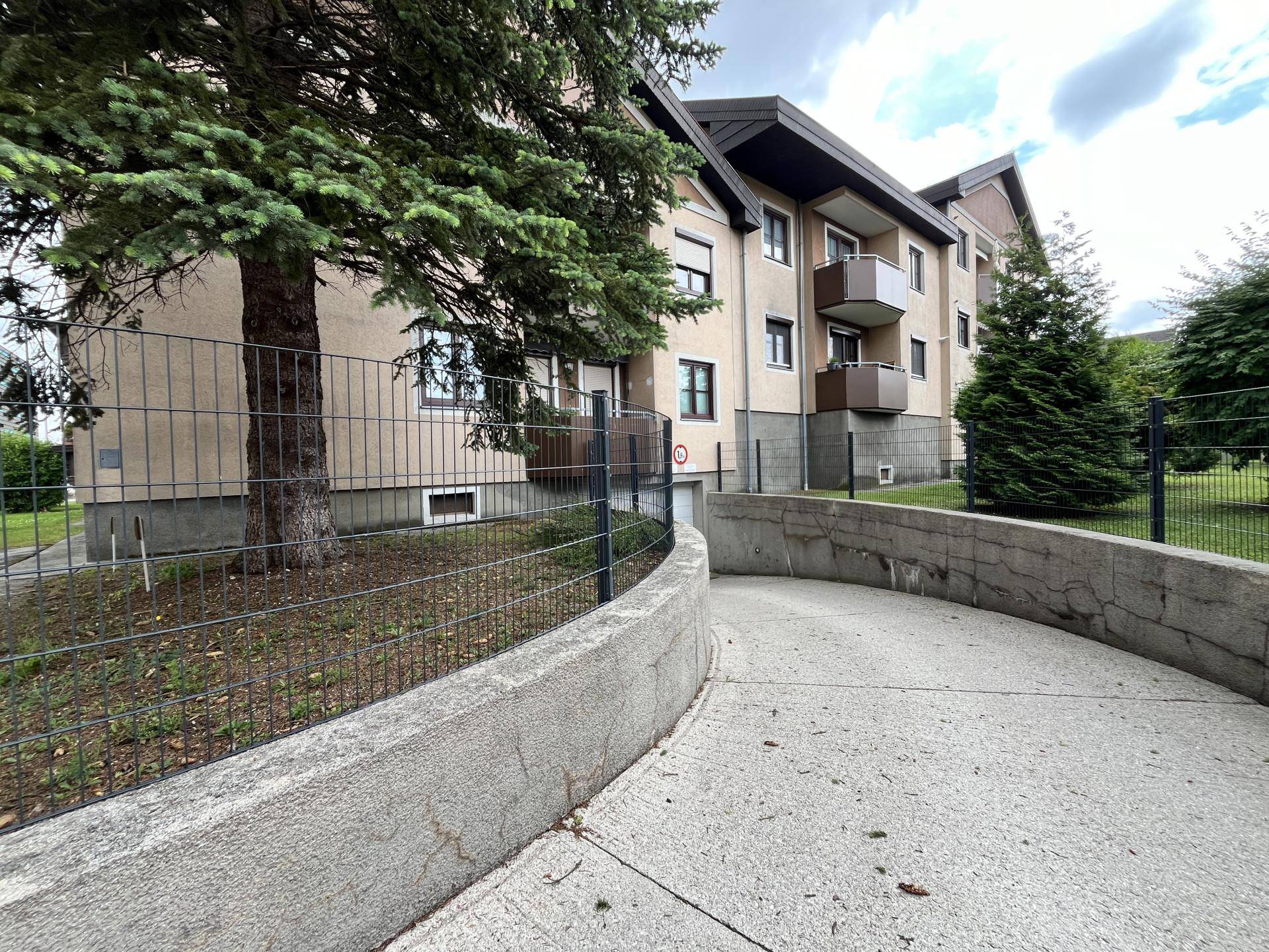 Mehrfamilienhaus mit Balkonen und einer Zufahrt zur Tiefgarage, umgeben von Bäumen und einer Grünfläche.