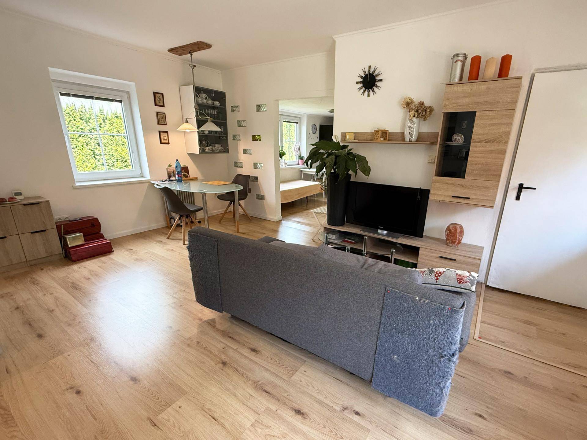 Heller Wohnbereich mit Sofa, Holzboden und Essbereich am Fenster mit Blick ins Grüne.
