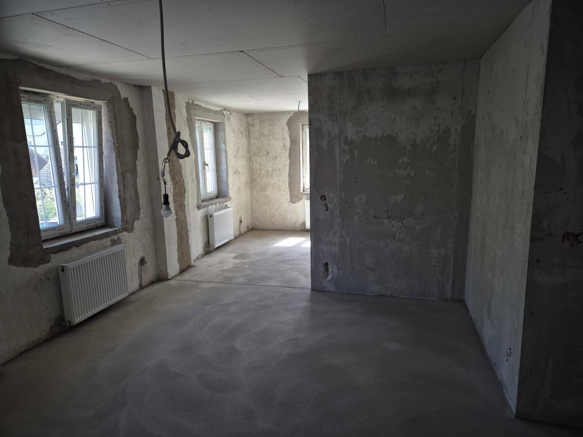 Unfertiger Innenraum mit Rohbauwänden, Betonboden und Fenstern, bereit für den Ausbau.
