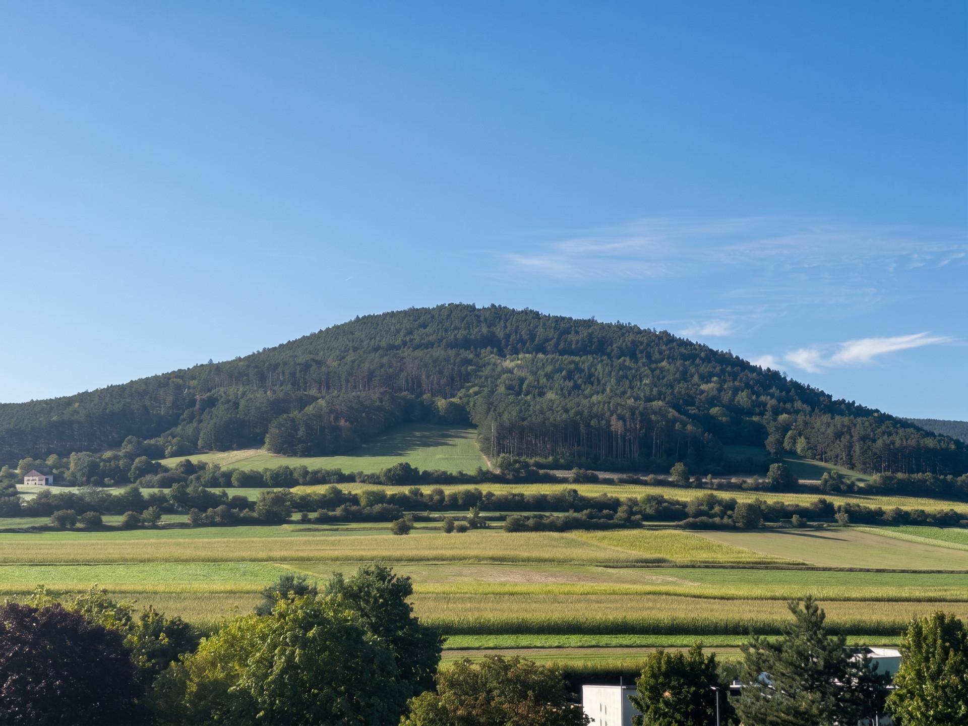 Malerische Landschaft mit bewaldeten Hügeln und grünen Feldern unter klarem blauen Himmel.