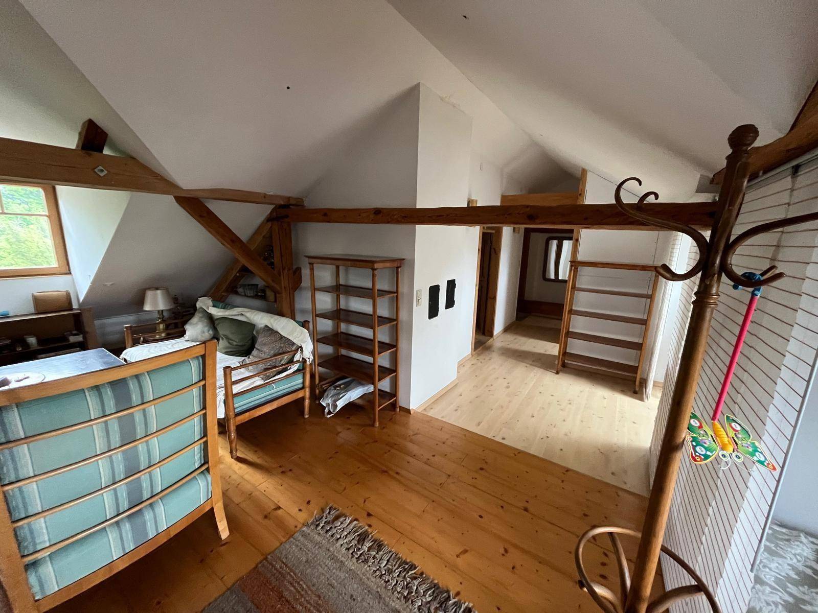 Geräumiges Dachzimmer mit Holzbalken, Holzboden und einfacher Einrichtung.