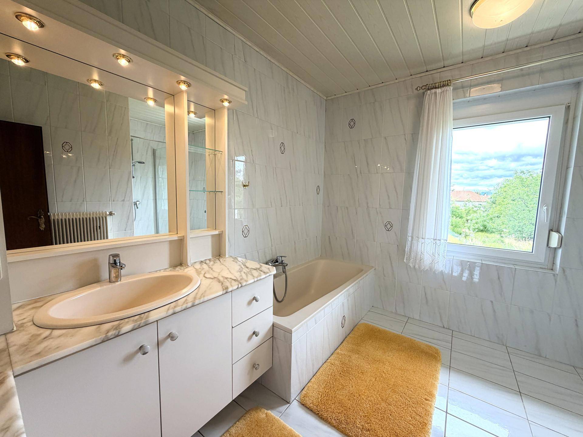 Helles Badezimmer mit Badewanne, Waschtisch und Fenster, komplett gefliest.