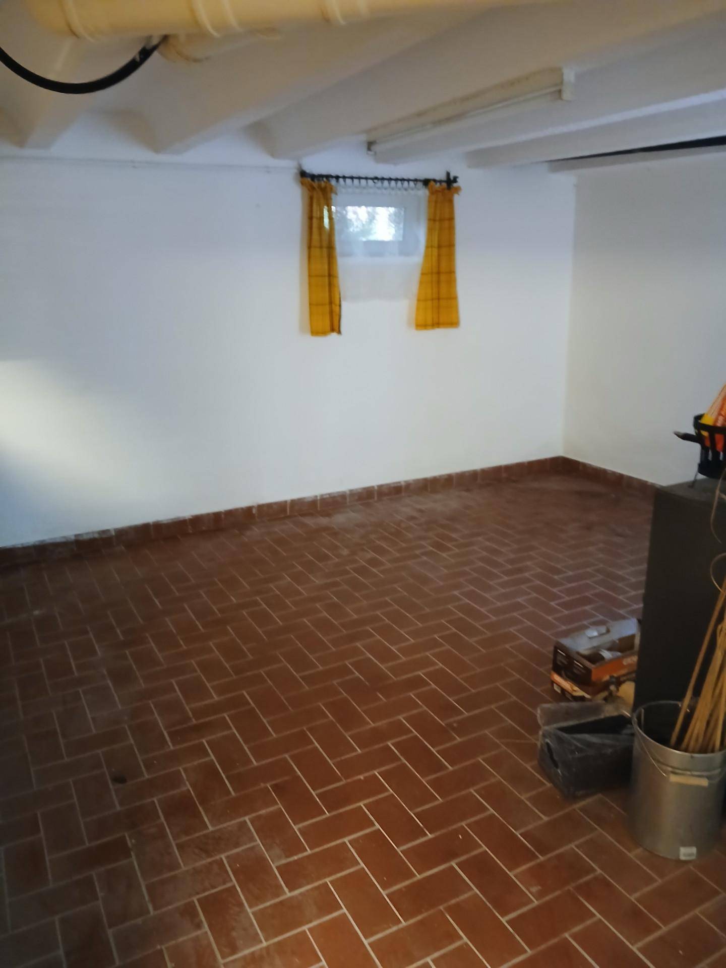 Kellerraum mit Terrakottafliesenboden, weiß gestrichenen Wänden und einem kleinen Fenster mit gelben Vorhängen.