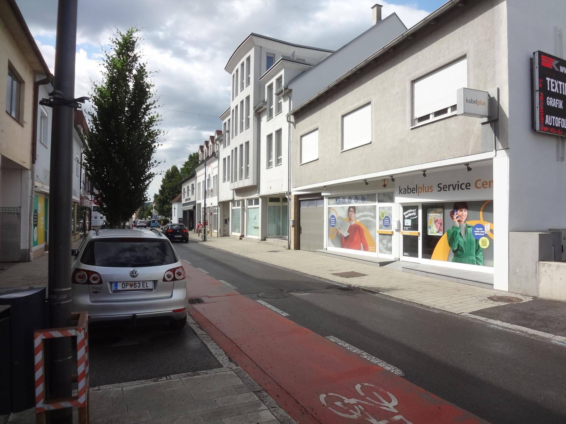 Belebte Straße mit geparktem Auto, Geschäftsgebäuden und einem Kabelplus Service Center mit modernen Schaufenstern.
