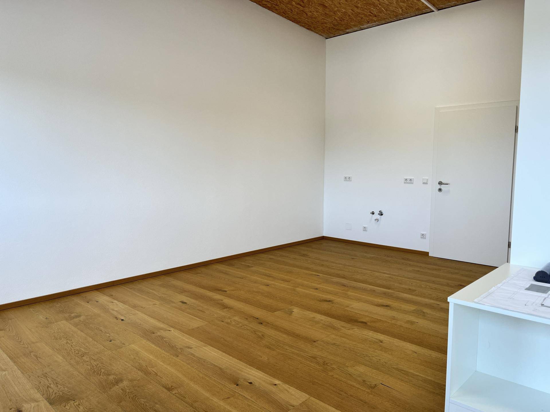 Ecke eines Raumes mit Holzboden, weißen Wänden und sichtbaren Anschlüssen für Strom und Wasser.
