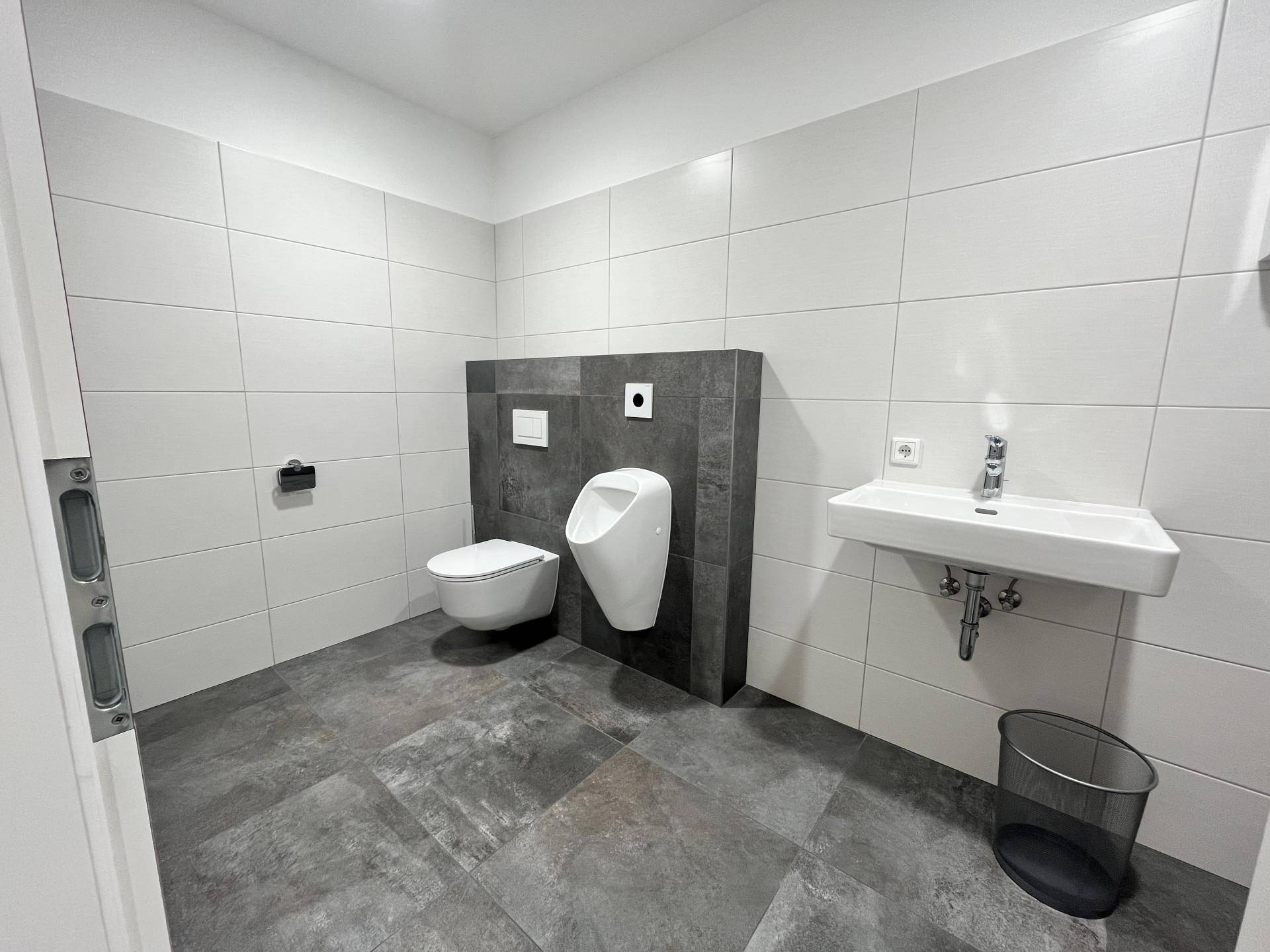 Modernes Badezimmer mit Urinal, Wand-WC und Waschbecken, komplett gefliest in Weiß und Dunkelgrau.