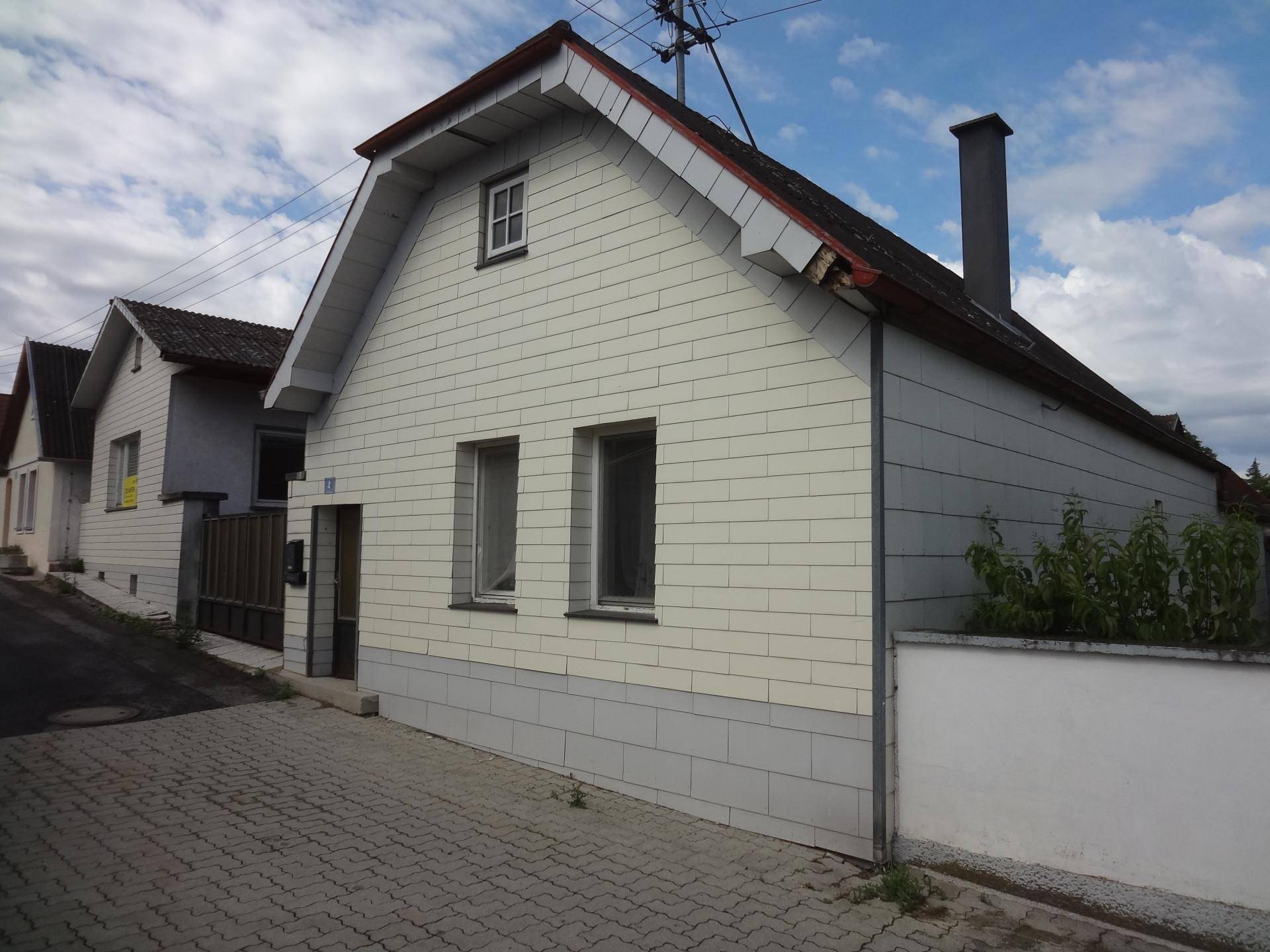 Ansicht des Hauses von der Straße mit weißer Fassade, braunem Dach und einem Schornstein.