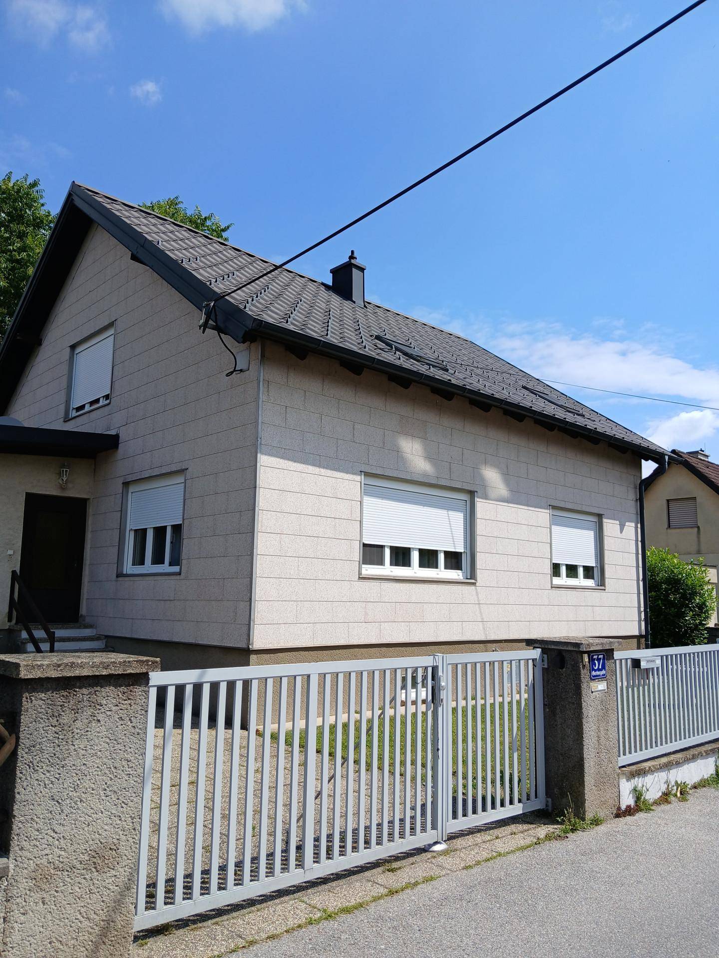 Seitenansicht des Hauses mit heller Fassade, Rollläden und einem Metallzaun zur Straße.