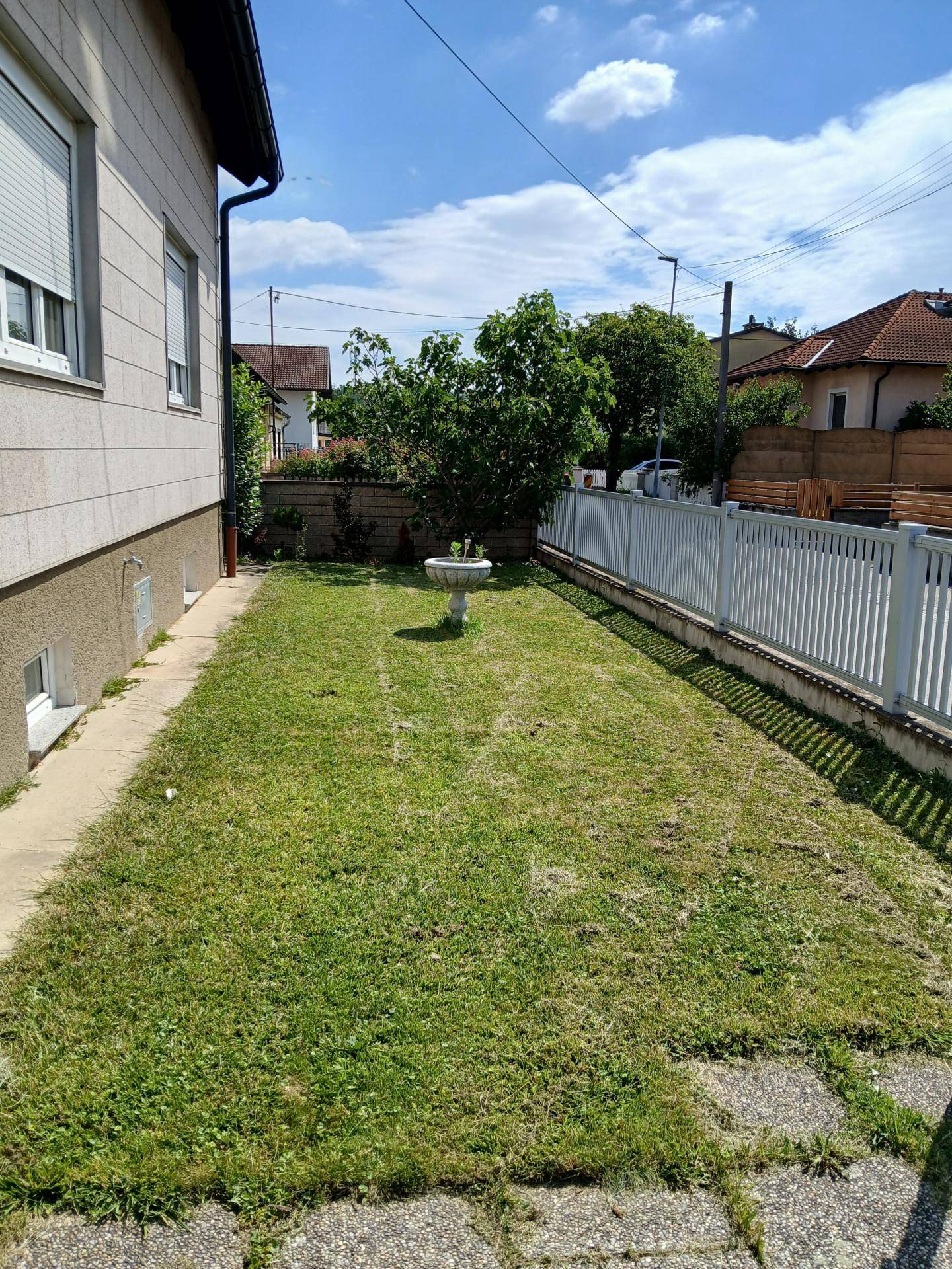 Schmaler Gartenstreifen neben dem Haus mit Rasenfläche, einem kleinen Brunnen und Zaun.