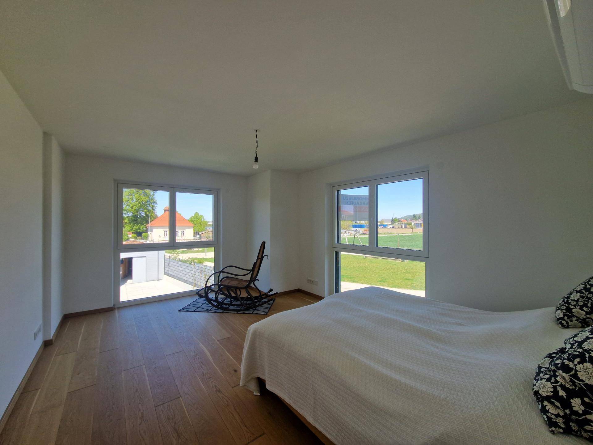 Helles Schlafzimmer mit Holzboden, großen Fenstern und Blick auf einen Spielplatz und die Umgebung.