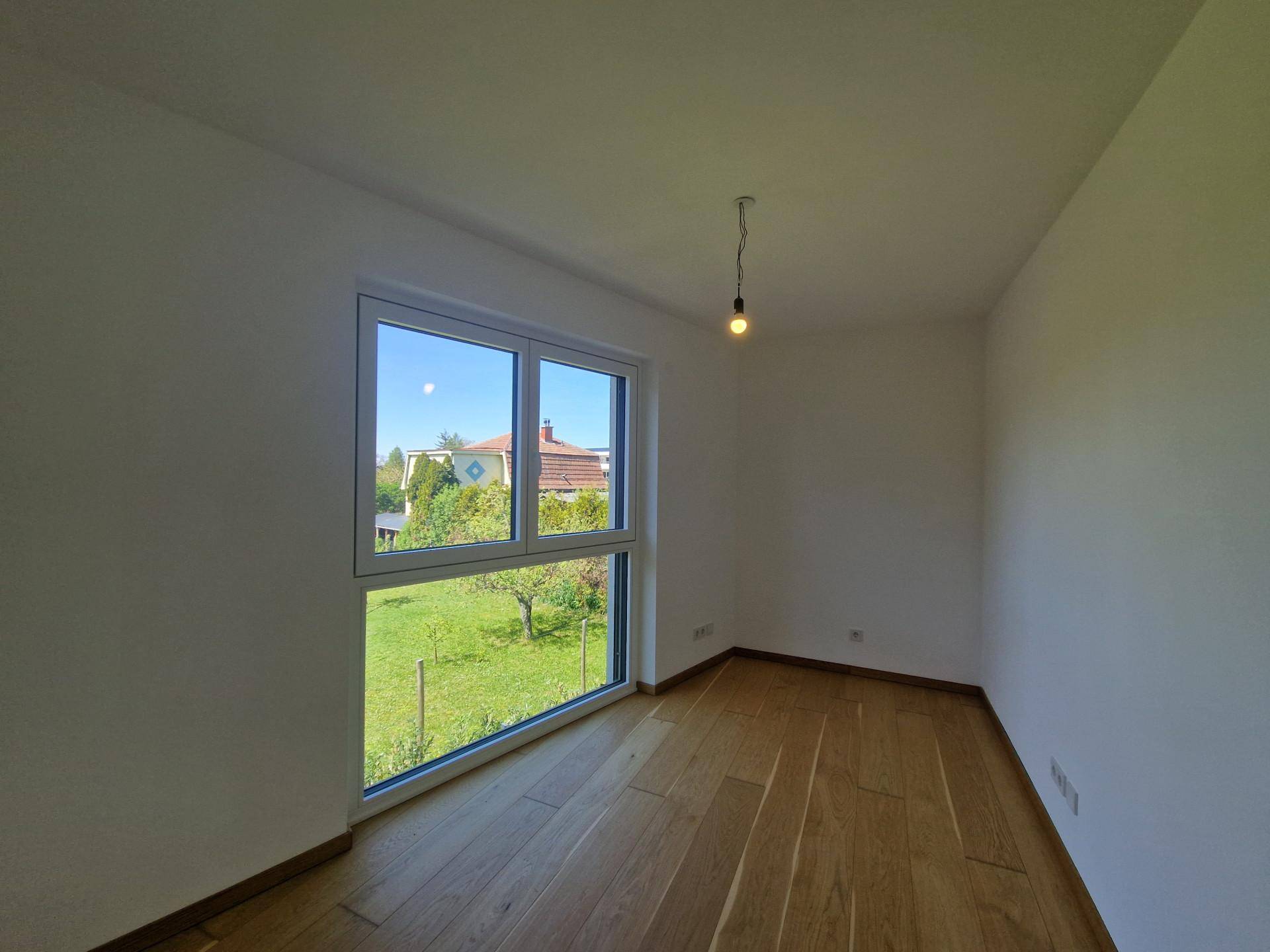Freundliches Zimmer mit Holzboden und großem Fenster, das einen schönen Blick in den grünen Garten ermöglicht.