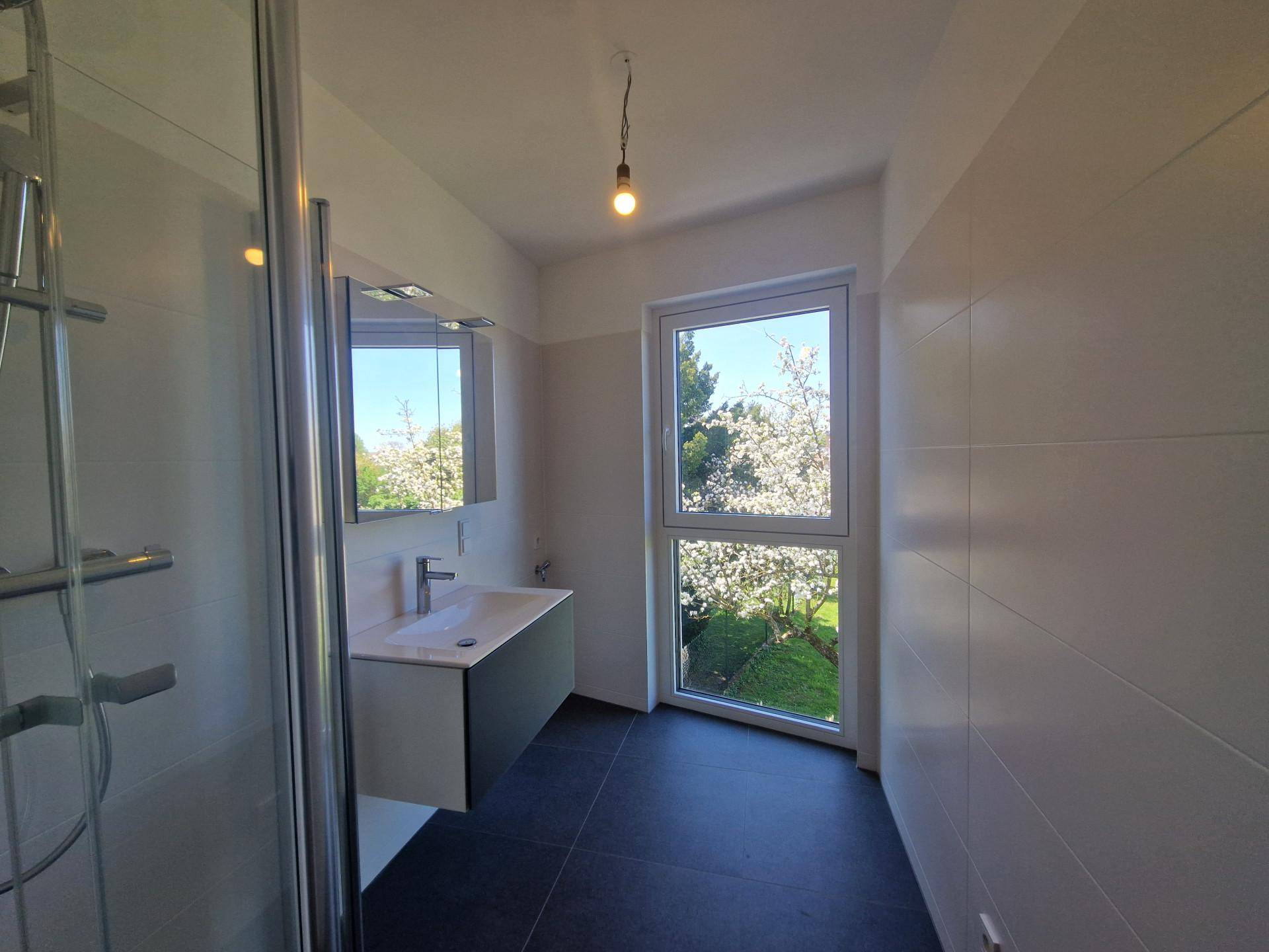 Modernes Badezimmer mit Duschkabine, Waschtisch und Fenster mit Blick auf einen blühenden Baum.
