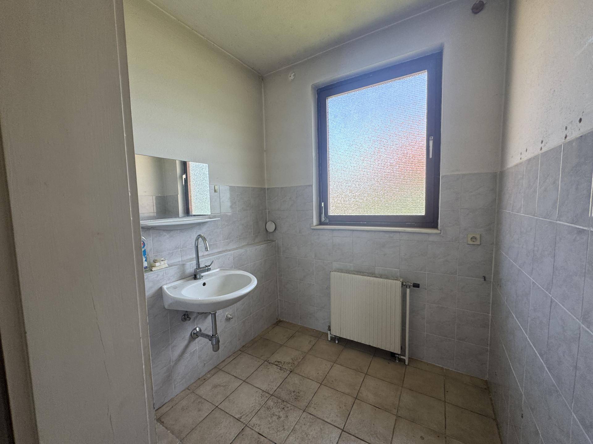 Badezimmer mit hellgrauen Fliesen, Waschbecken, Spiegel und einem Fenster.