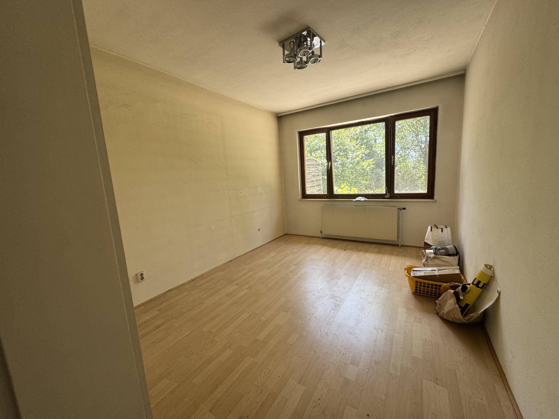 Helles, leeres Zimmer mit Parkettboden, großen Fenstern und einem Heizkörper.