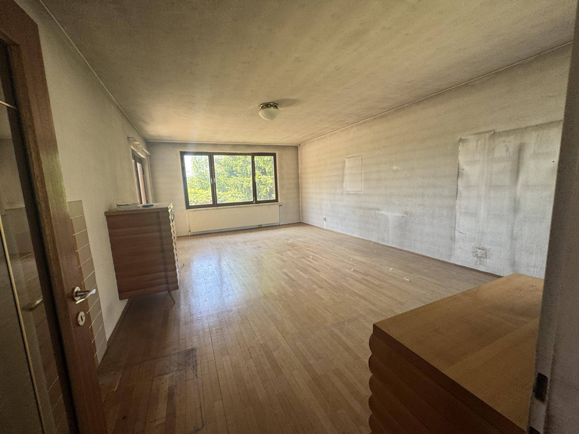 Leeres Zimmer mit Parkettboden, großen Fenstern und Heizkörper, bereit zur Renovierung.