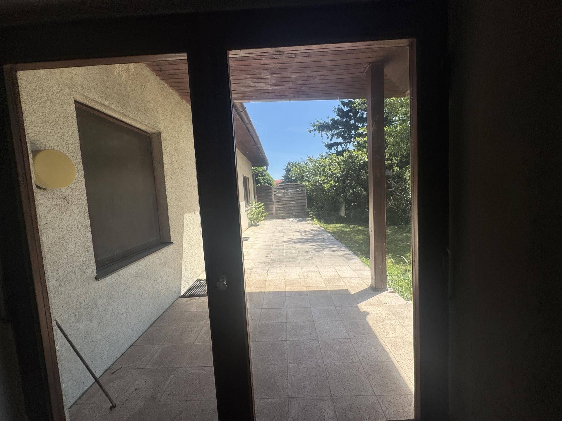 Blick durch eine Glastür auf eine gepflasterte, überdachte Terrasse und den Garten.