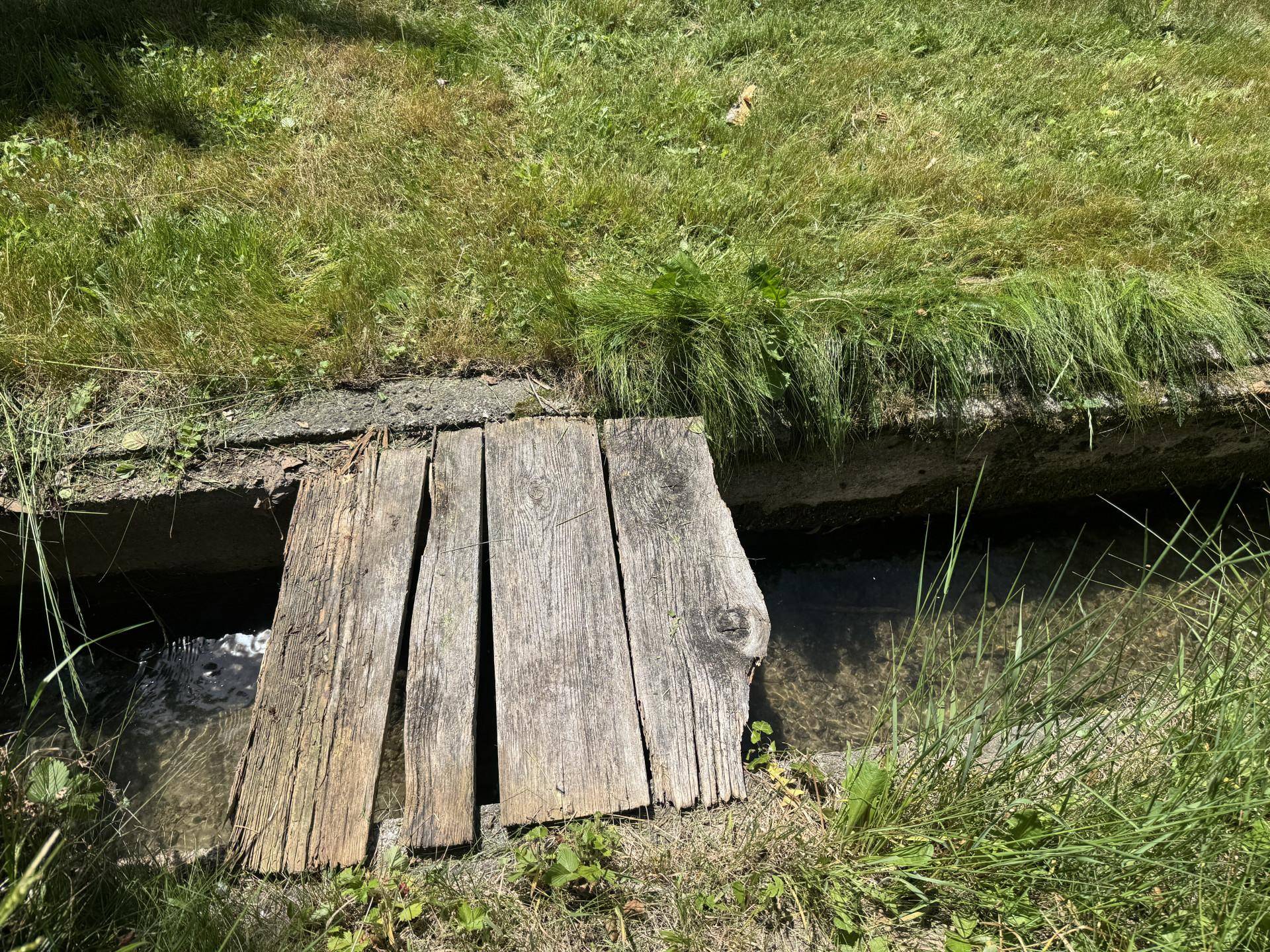 Kleine Holzbrücke über einen schmalen Bach oder Graben in einer grünen Wiese.