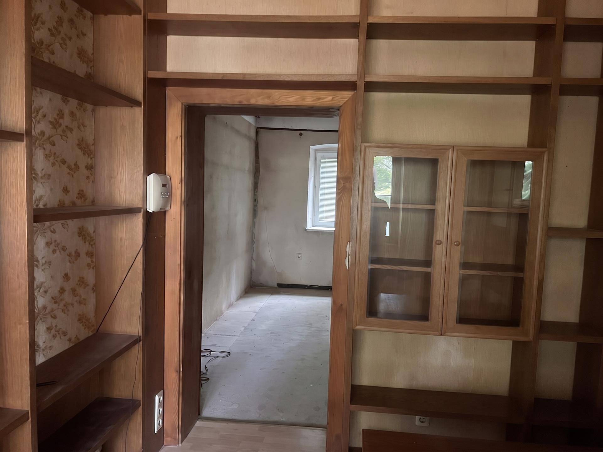 Innenraum mit umfangreichen Holzregalen und einem Durchgang zu einem weiteren Zimmer mit Fenster.