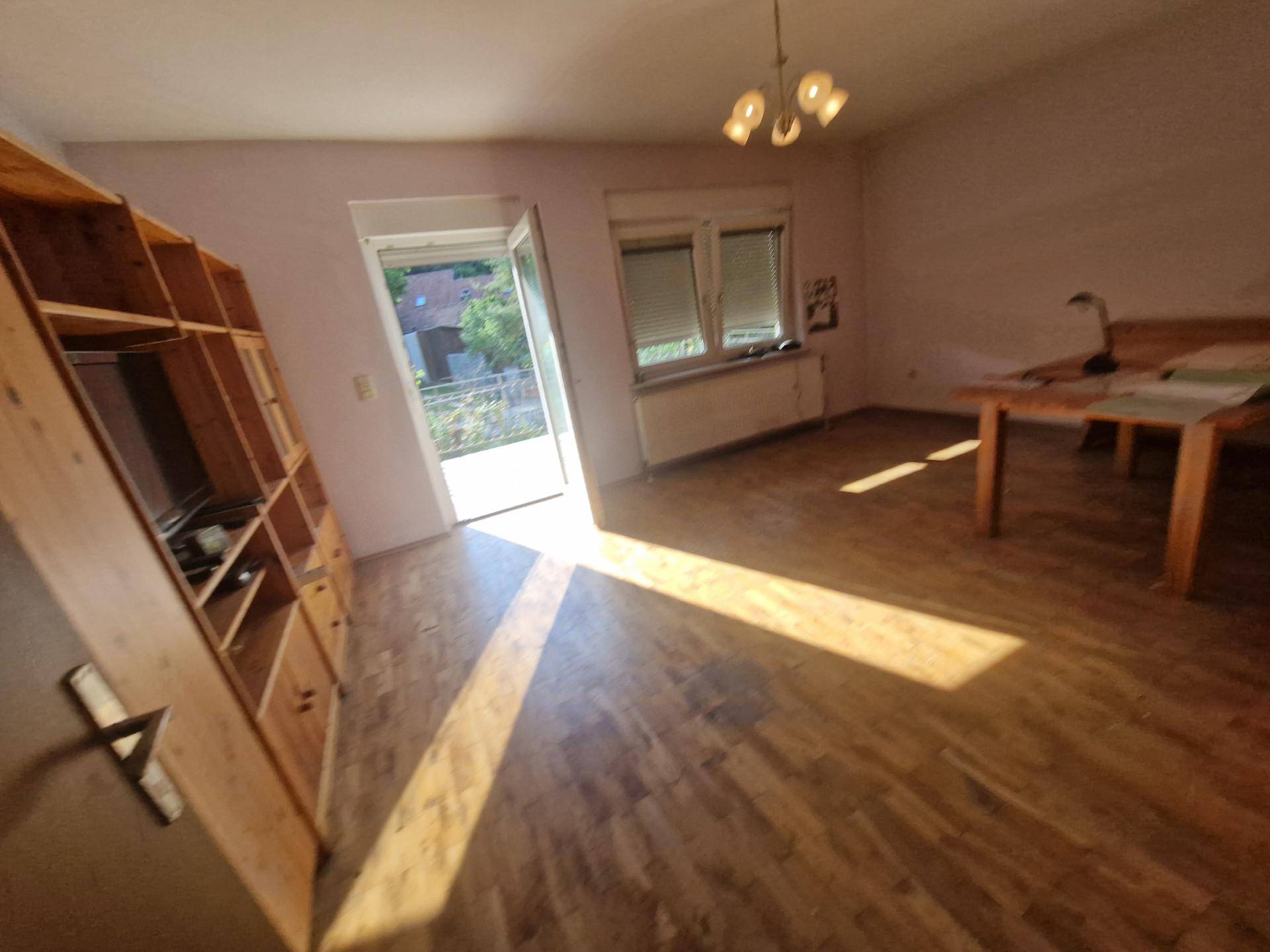 Helles Wohnzimmer mit Holzregal, Tisch und Zugang zum Balkon.
